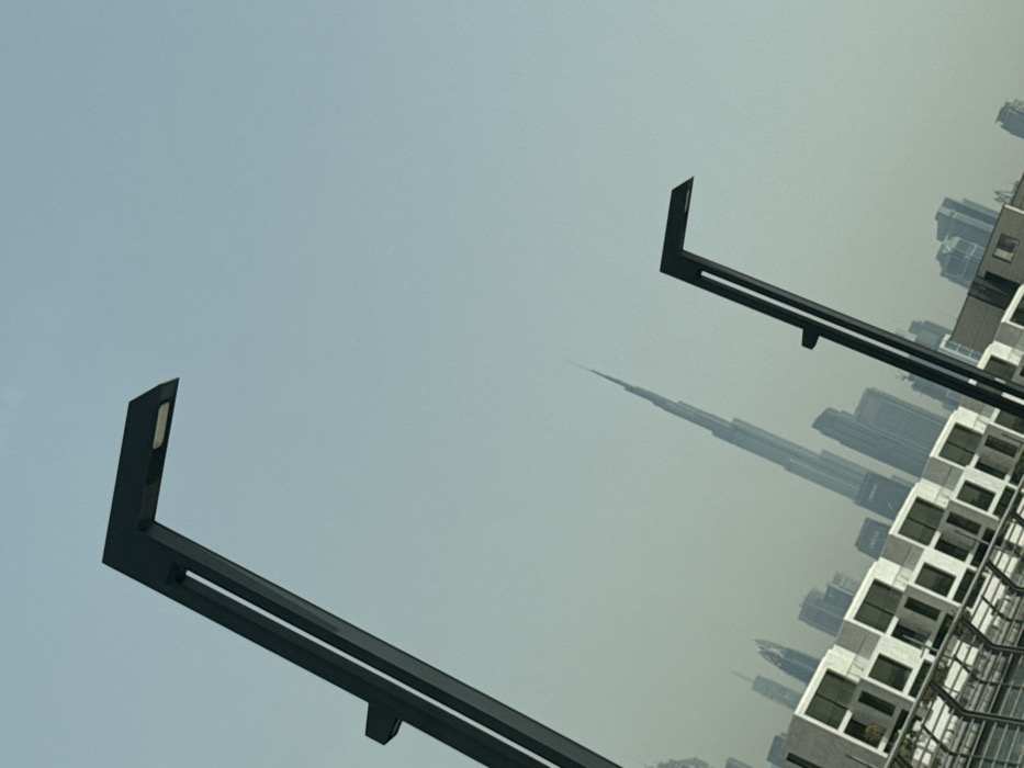 The image displays an urban scene in Dubai, United Arab Emirates, captured from a low-angle perspective with a significant tilt. In the immediate foreground, two modern, dark-colored streetlights with angled, L-shaped tops and integrated light panels ascend diagonally from the lower corners of the frame. The left streetlight is largely visible, while the right one is partially cropped. Behind these, a row of multi-story, light-colored buildings with numerous square window sections is visible, below which a metallic fence or railing structure runs horizontally. Dominating the background is the distinctively spired silhouette of the Burj Khalifa skyscraper, appearing hazy against the pale blue sky, flanked by other smaller high-rise buildings that form a city skyline receding into the atmospheric haze. No individuals, specific actions, or direct interactions are visible.