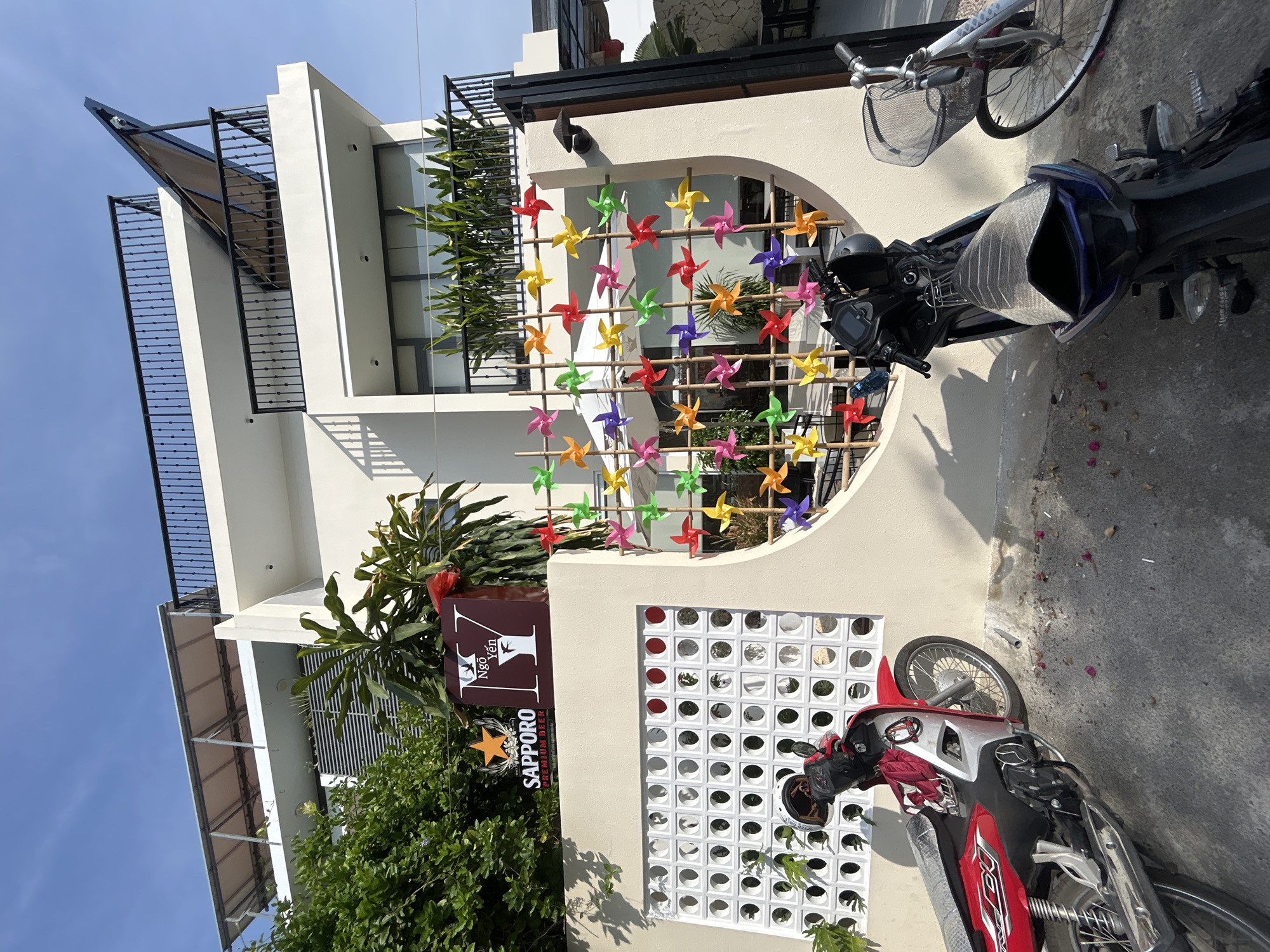 The image captures a street view in Nam Nha Trang Ward, Vietnam, on a sunny day with a clear blue sky. The foreground features three parked vehicles: a red and silver motorcycle on the left, a dark blue motorcycle in the center, and a bicycle with a wire basket on the right. The motorcycles are positioned on a concrete surface littered with fallen petals.
Behind the vehicles, a pale cream-colored wall forms the base of a modern building. The wall is adorned with a decorative circular motif filled with numerous colorful pinwheels of varying shades of red, orange, yellow, green, blue, and purple, attached to bamboo poles. To the left of the pinwheels is a white decorative lattice screen.
Above the wall, the building is a multi-story structure with white exterior walls and dark metal railings on balconies. The upper floors have large windows and awnings. A prominent sign with the SAPPHORO PREMIUM BEER logo and another sign with Vietnamese text that appears to say "Ngo Yen" are affixed to the wall, near some greenery. The overall scene suggests a commercial establishment or residential building with a decorative facade.
