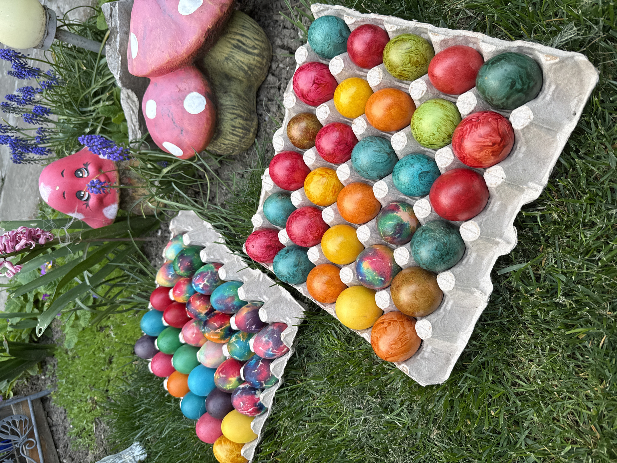 Two egg cartons filled with dyed Easter eggs sit on a patch of green grass. A whimsical garden arrangement is nearby, featuring a small red mushroom-shaped face, painted mushrooms, and flowering plants. The location may be Lukovit, Bulgaria.