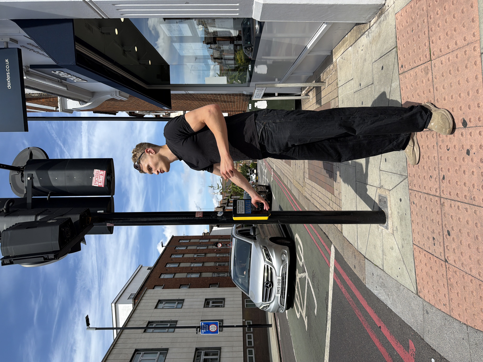 This media file captures a scene on a sunny day in London, United Kingdom. The primary subject is a young man, standing on a sidewalk and reaching out to press a pedestrian crossing button. He is dressed casually in a black t-shirt, black jeans, and light-colored shoes. The man appears focused on the task of crossing the street.
The setting is an urban environment with a street, sidewalk, and buildings. To the left, a building with a dark awning and large windows is visible, with a sign displaying "dexters.co.uk". Across the street, a silver Mercedes-Benz van is stopped at the pedestrian crossing. The street is marked with red and white lines, indicating a crosswalk and a cycle lane. In the background, residential buildings with multiple windows can be seen.
The time of day is likely daytime, evidenced by the clear blue sky with wispy clouds and the strong shadows cast on the sidewalk. The weather is pleasant, with sunshine illuminating the scene. The presence of a pedestrian crossing, the style of the buildings, and the license plate on the van (BM19 HBB) are all consistent with a UK location. The overall mood is one of everyday urban activity, with the man engaging in the common act of crossing a street. Visible text includes "dexters.co.uk", "PEDESTRIANS", "WAIT", "for signal appeals", and a sign with a blue circle containing a white pictogram of a bicycle and the number "3.5m".