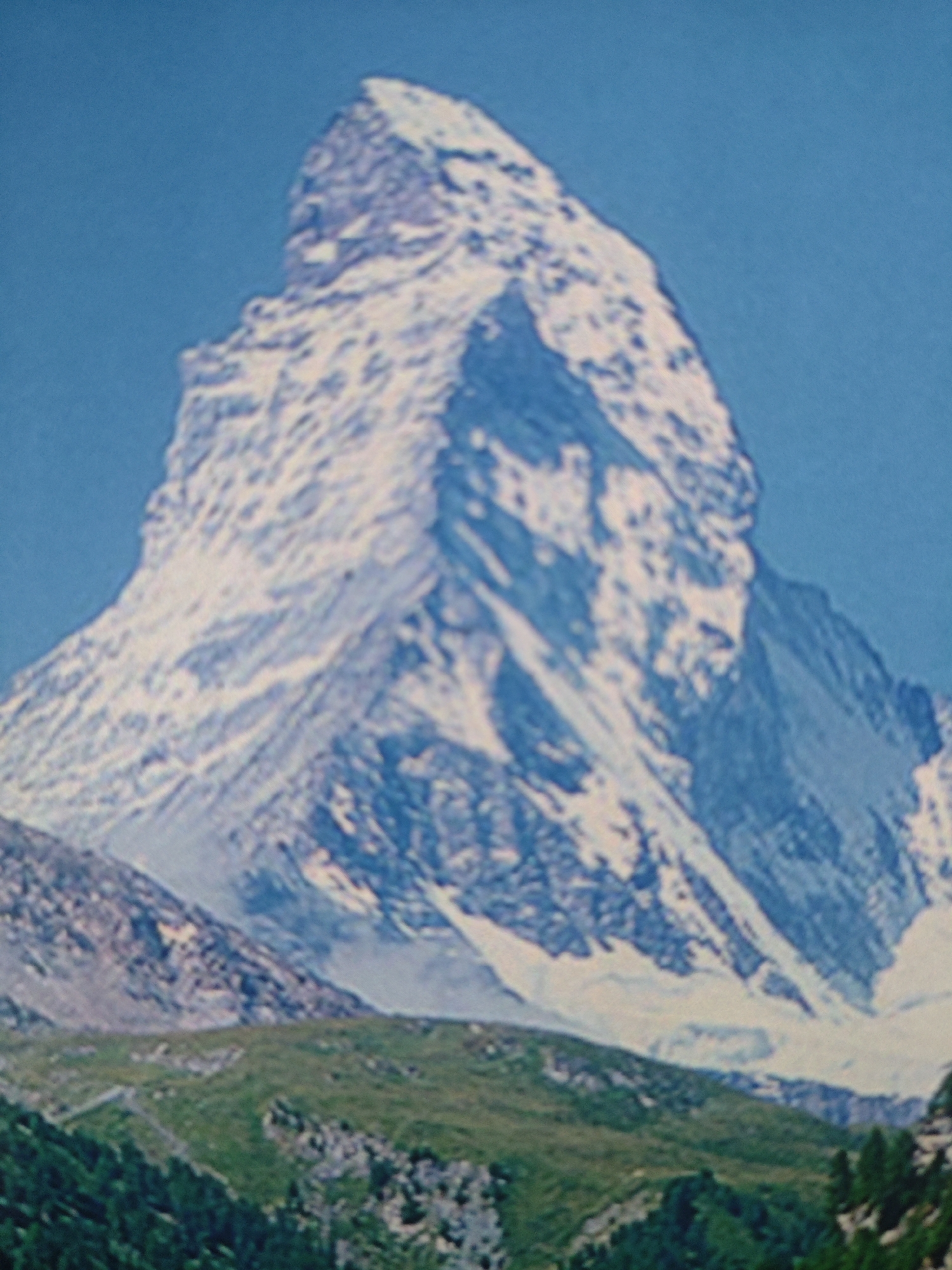 This is a close-up, low-resolution, color photograph of the Matterhorn mountain, a jagged peak in the Alps. The mountain is snow-capped and appears to be made of rock and ice. The sky is a clear blue. In the foreground, there is a green, grassy slope with some trees at the bottom. The image is out of focus and appears to be a postcard or a vintage photograph. It is not possible to determine the location as Krasnaya Polyana, Russia, as the Matterhorn is in Switzerland.