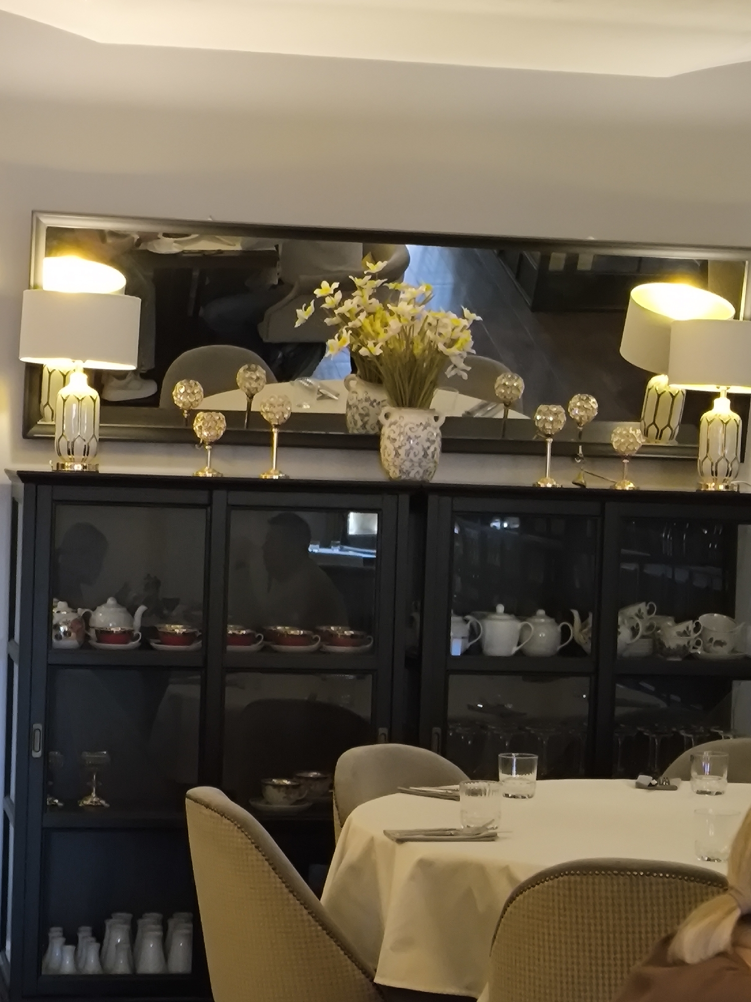 The image depicts a dining area, likely within a restaurant or hotel, possibly located in Smolensk, Russia. A table with a white tablecloth is surrounded by upholstered chairs. The table is set with glassware and silverware, indicating that it is ready for service.
Behind the table is a dark-colored cabinet or sideboard with glass doors, displaying a collection of teacups, saucers, teapots, and other decorative items. The top of the cabinet is adorned with two lamps, decorative spheres on stems, and a vase filled with flowers. A large mirror hangs above the cabinet, reflecting the surroundings and adding depth to the scene. The lighting appears to be soft and ambient, creating a warm and inviting atmosphere.