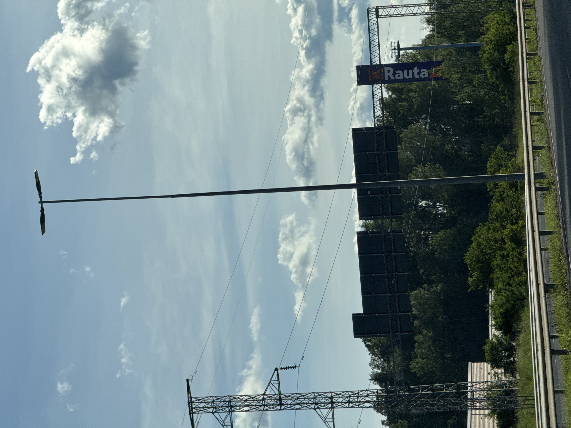 An outdoor daytime shot captures a tall street lamp and electrical pylons against a blue sky with scattered white clouds. A dense line of green trees forms the background, with a highway barrier in the foreground. A vertical blue banner with white lettering reading "Rauta" is visible on the right side of the image. There are no people or animals visible in the scene. The setting appears to be an urban or suburban environment, possibly near a commercial area given the presence of signage.