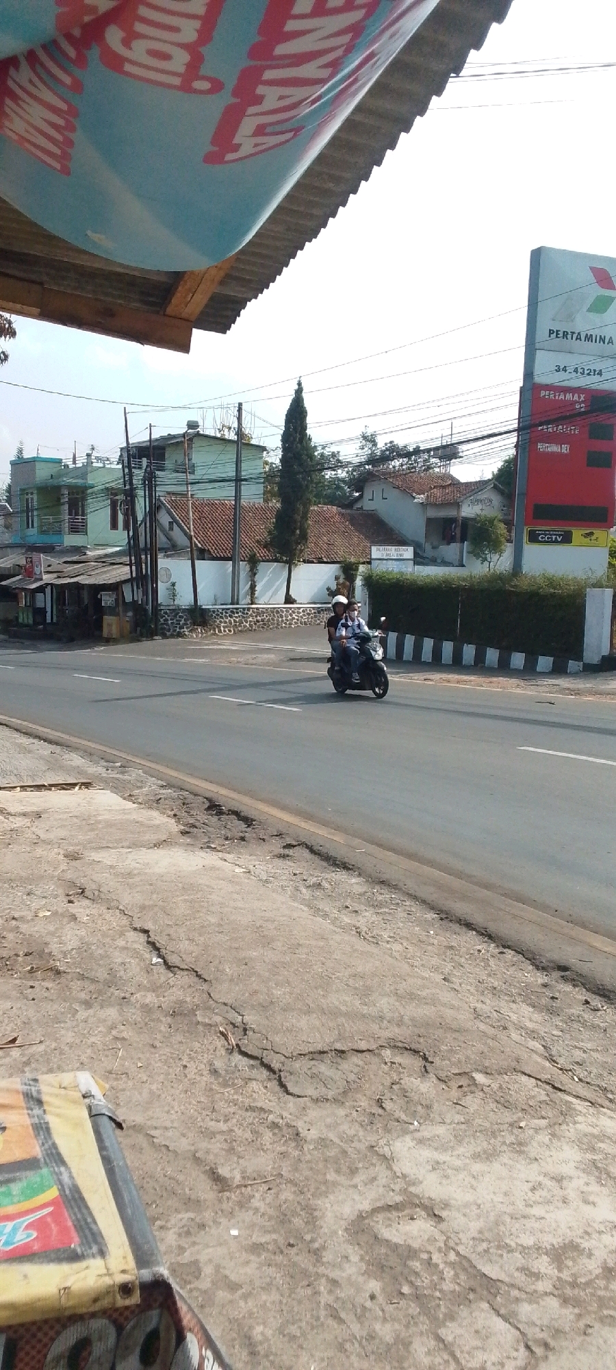 In Cilaku, Indonesia, a motorcycle with two occupants, a driver and a passenger, rides on a paved road. The driver wears a white helmet and a light-colored shirt, while the passenger has a white helmet and a blue shirt. The road is lined with a grassy hedge and a white fence with black vertical stripes on the right side. Behind the hedge, a tall, slender tree stands out against the backdrop of houses and utility poles. To the right of the tree, a red sign with white text indicates "PERTAMAX" and other fuel prices, along with a "CCTV" warning sign. A large, partially visible banner with blue and red lettering is suspended above a corrugated metal roof on the left side of the image. The foreground features cracked and worn concrete.