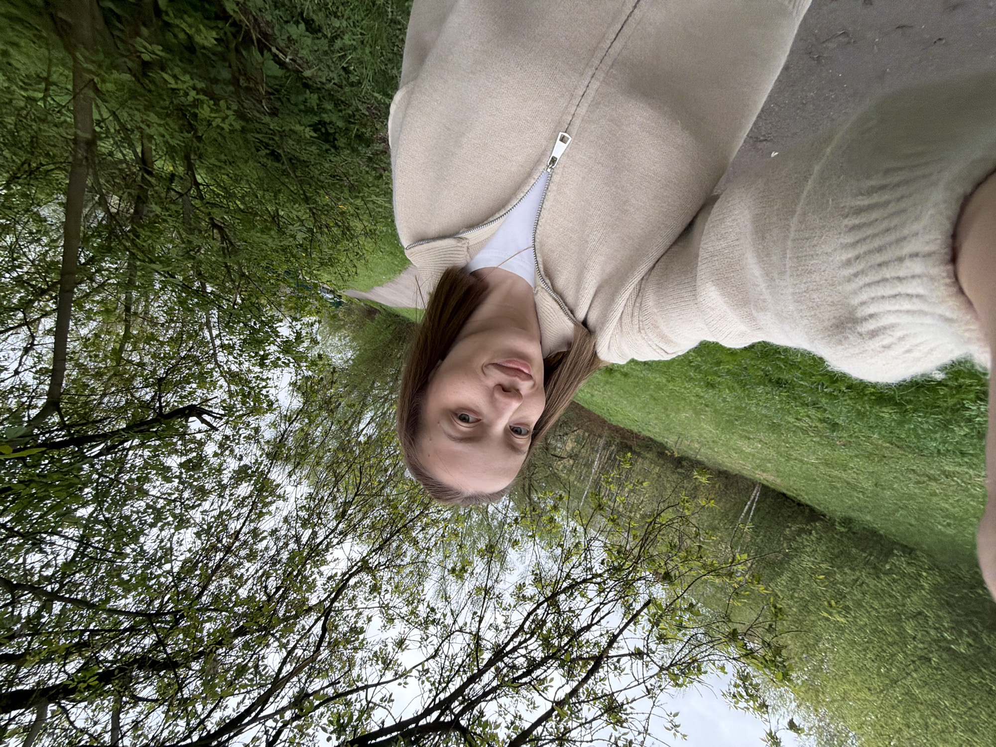 A woman is taking a selfie outdoors in Reshetnikovo, Russia. She is wearing a beige cardigan and a white shirt. The background shows a path flanked by green grass and trees, suggesting a park or a nature area. The sky is visible through the branches of the trees.