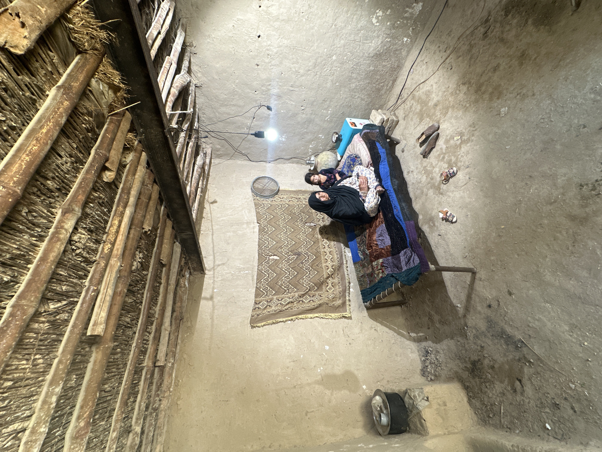 A high-angle indoor shot depicts a scene in what appears to be a modest dwelling in Quetta City Tehsil, Pakistan. The room has rough, unadorned walls, possibly made of mud or clay, and a simple ceiling with a single bare light bulb hanging from exposed wires.
In the center of the room, an adult female wearing a dark hijab and patterned clothing is lying on a bed with a child next to her. The bed is covered with colorful quilts and blankets.
Near the bed, there is a patterned rug on the floor, a metal fan, and several pairs of sandals.
The left side of the frame shows a wall constructed with bamboo or similar natural materials, indicating a rudimentary construction method. There is an aluminum pan/pot on the floor in the lower right. The room appears to be dimly lit and sparsely furnished, suggesting a scene of simple living.