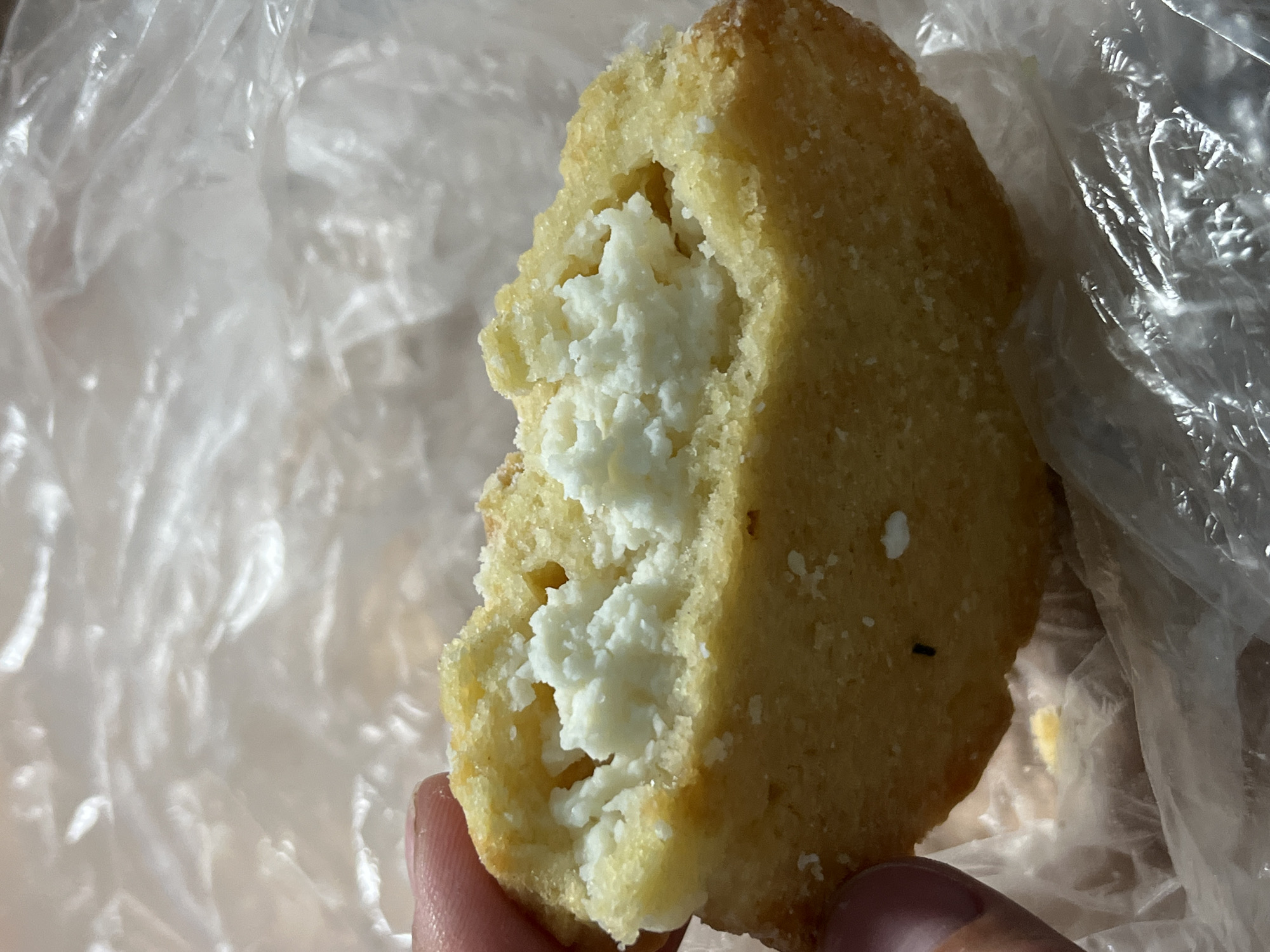 A close-up, slightly overhead shot shows a person's thumb and index finger holding a partially eaten, round, golden-brown pastry with a white, crumbly filling. The pastry is broken open, revealing the creamy white interior. The background consists of crinkled, transparent plastic material. There is no discernible information about the location in Nizhny Tagil, Russia.