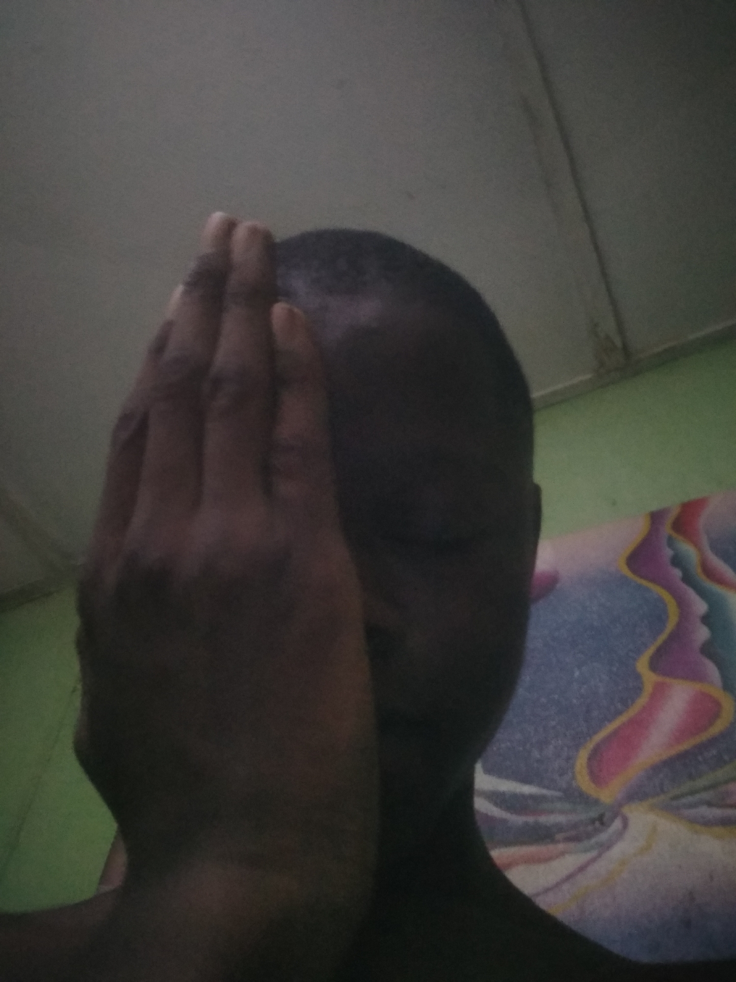 A close-up, dimly lit, low-angle indoor shot features a person with dark skin covering their face with their right hand. The person has very short, dark hair. Their eyes and most of their facial features are obscured by the hand. The activity suggests a moment of shyness, concealment, or perhaps a pensive mood, but no specific emotion is discernible.
The background consists of a light-colored ceiling with a visible seam or line towards the top. A muted green wall or surface is visible on the left side of the frame. To the right of the person, a vibrant, abstractly patterned textile is present, likely a blanket, sheet, or tapestry. This textile features flowing, organic shapes and patterns in shades of purple, pink, yellow, and blue, with some designs vaguely resembling overlapping profiles of faces or abstract ribbons. There is no visible text in the image. The low light suggests an indoor setting either in the evening or a room with limited natural light.