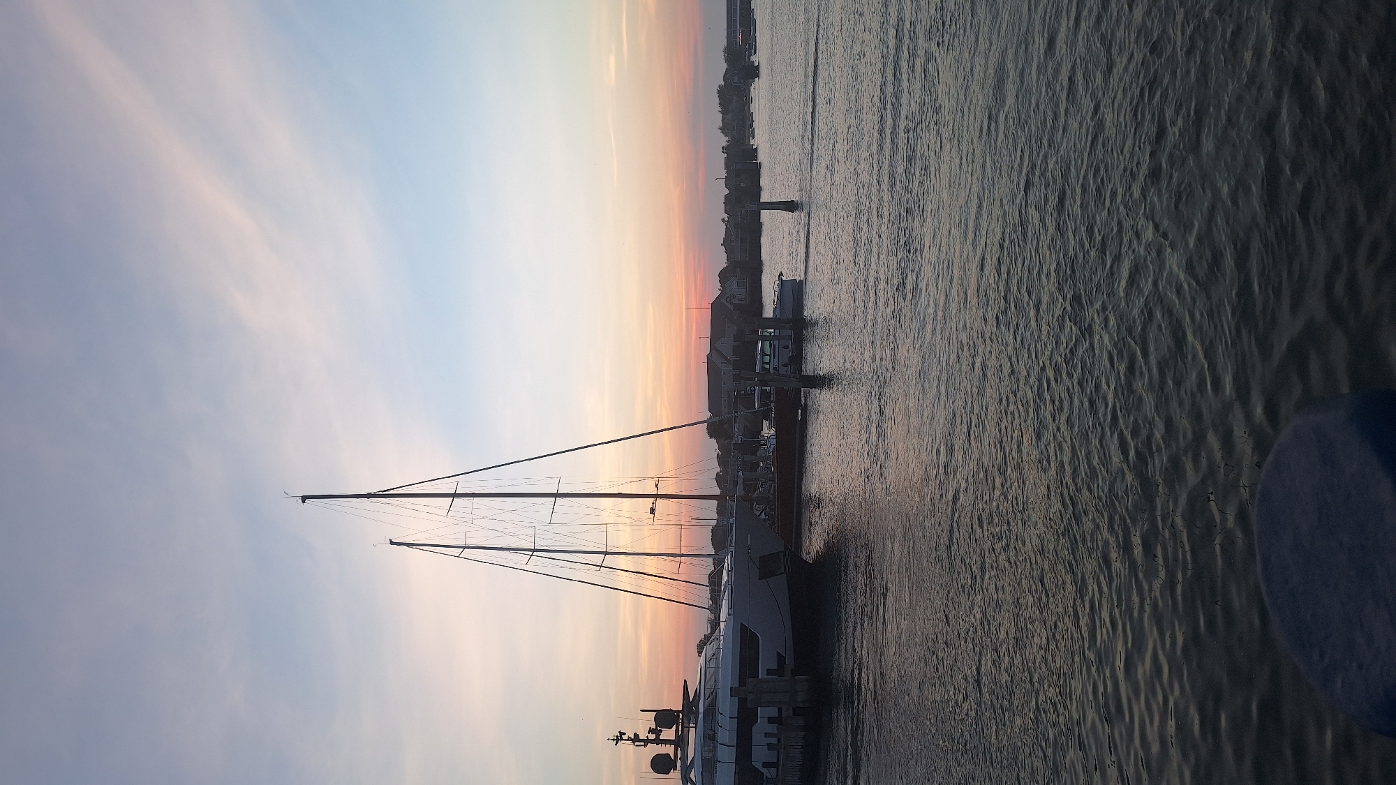 The image depicts a harbor scene at sunset in Nantucket, Massachusetts, United States of America. Dominating the foreground is the stern of a large white yacht, moored to a dock. To its left, a smaller, darker boat is visible. In the mid-ground, several sailboats with tall masts are docked, their rigging stark against the colorful sky. The water in the harbor is calm, reflecting the hues of the sunset. In the background, a row of buildings lines the shore, suggesting a residential or commercial area. The sky is a gradient of soft pinks, oranges, and blues, with thin, wispy clouds. The overall impression is one of tranquility and a picturesque evening in a coastal town.