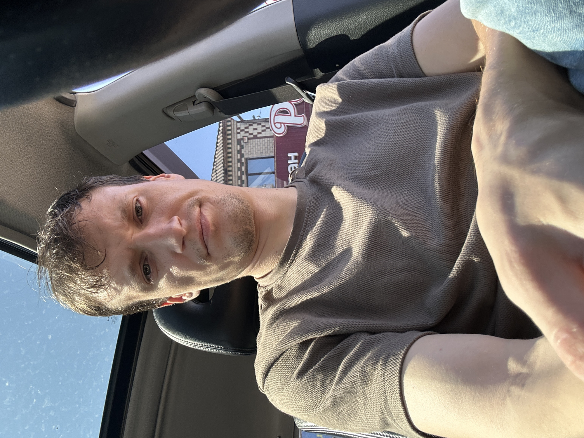A man with short, brown hair and stubble is sitting in the driver's seat of a car. He is wearing a brown, waffle-knit t-shirt. The car has a sunroof open, revealing a clear blue sky with some dust motes. Through the car window, a building with decorative brickwork and a sign with red lettering and a white "P" is visible. The man is looking directly at the camera with a neutral expression. The lighting suggests it is daytime.