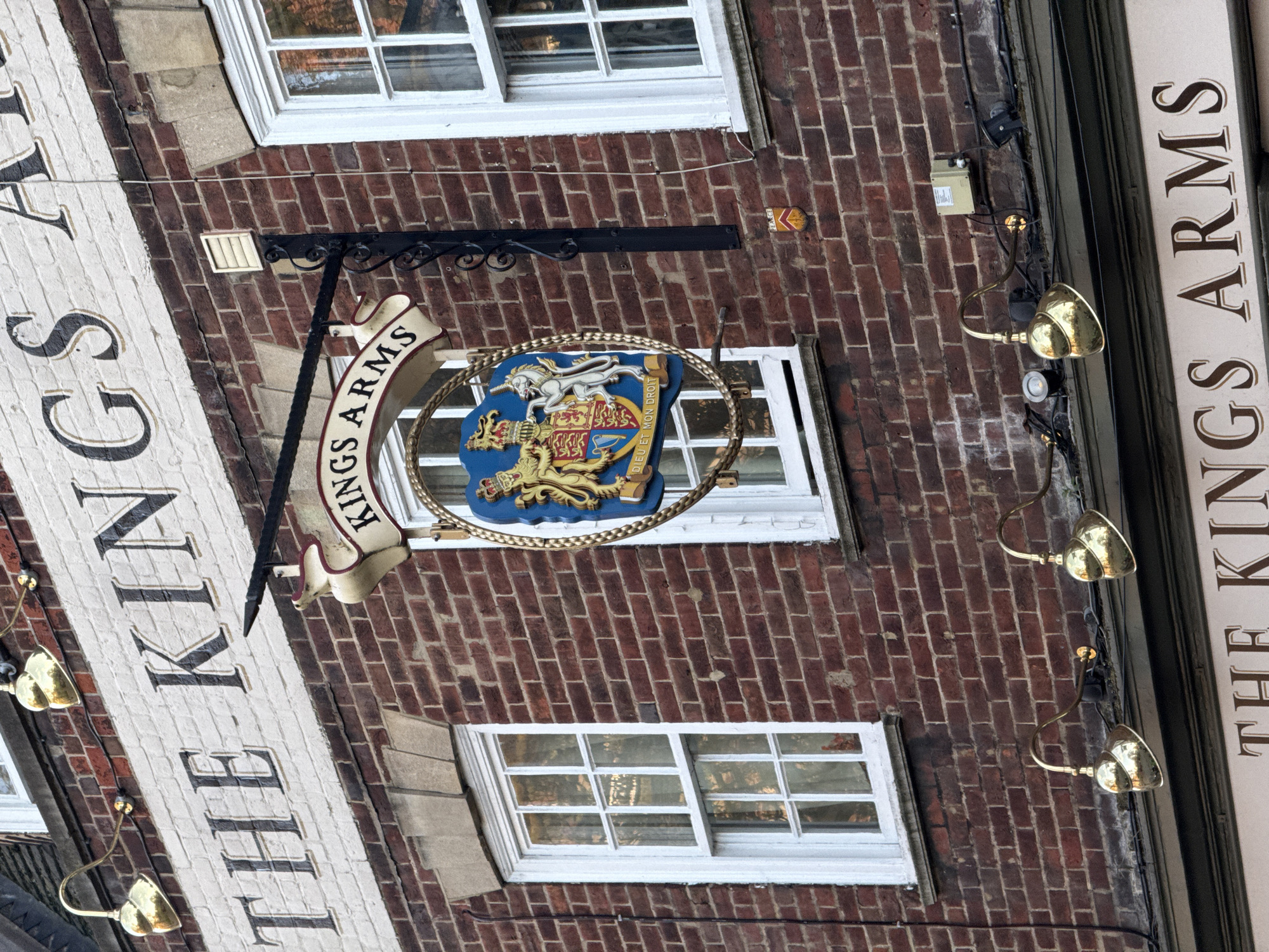 The image depicts the exterior of "The King's Arms" pub, likely located in London, United Kingdom. The pub sign features the words "THE KING'S ARMS" vertically on a white background, and a separate sign that hangs above the window includes the text "KING'S ARMS" in an arc shape. This sign includes the British royal coat of arms. The building has a red brick facade and windows with white frames. Golden lamps illuminate the building.