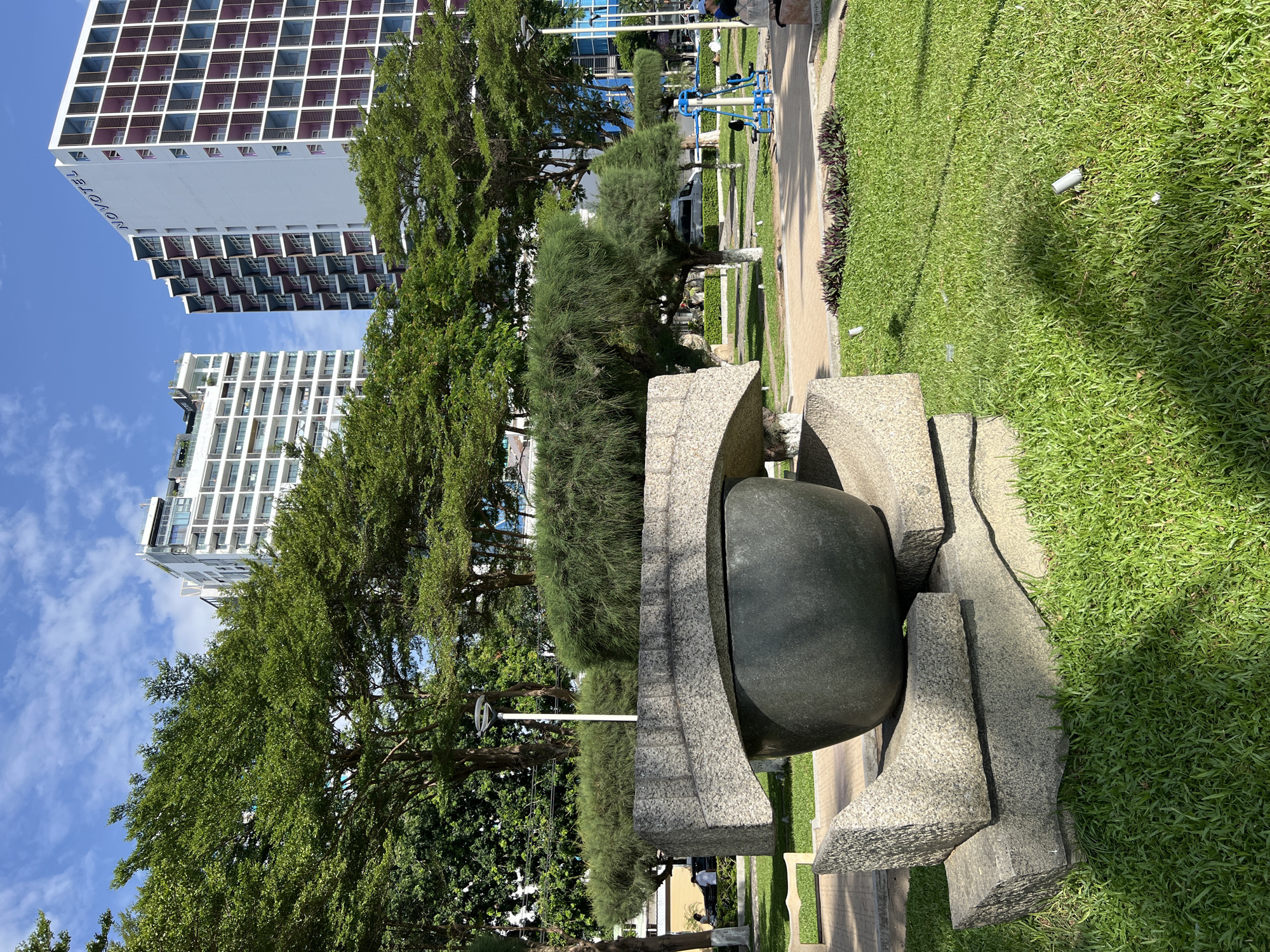 This image depicts an outdoor scene in Nha Trang, Vietnam, featuring a contemporary stone sculpture in the foreground. The sculpture consists of rough-hewn granite blocks supporting a large, dark, bowl-shaped object, possibly a basin or an abstract representation. Behind the sculpture, a meticulously manicured lawn transitions into paved pathways and decorative plant beds.
In the mid-ground, lush green trees with feathery foliage are interspersed with modern high-rise buildings. One building is clearly identifiable as the Novotel Nha Trang, marked by signage and its distinctive grid-like facade with purple accents. Another adjacent white building also exhibits a contemporary architectural style with balconies. A street lamp with a circular top is visible among the trees. Further in the background, a blue outdoor exercise machine is situated on the grass, and a few individuals are present, one possibly sitting on a bench. A light-colored van is parked on a road behind the exercise equipment. The sky above is a vibrant blue with scattered white clouds, suggesting a clear and sunny day. The overall environment appears to be a well-maintained public space or park area, likely a tourist-friendly location given the presence of the Novotel hotel.