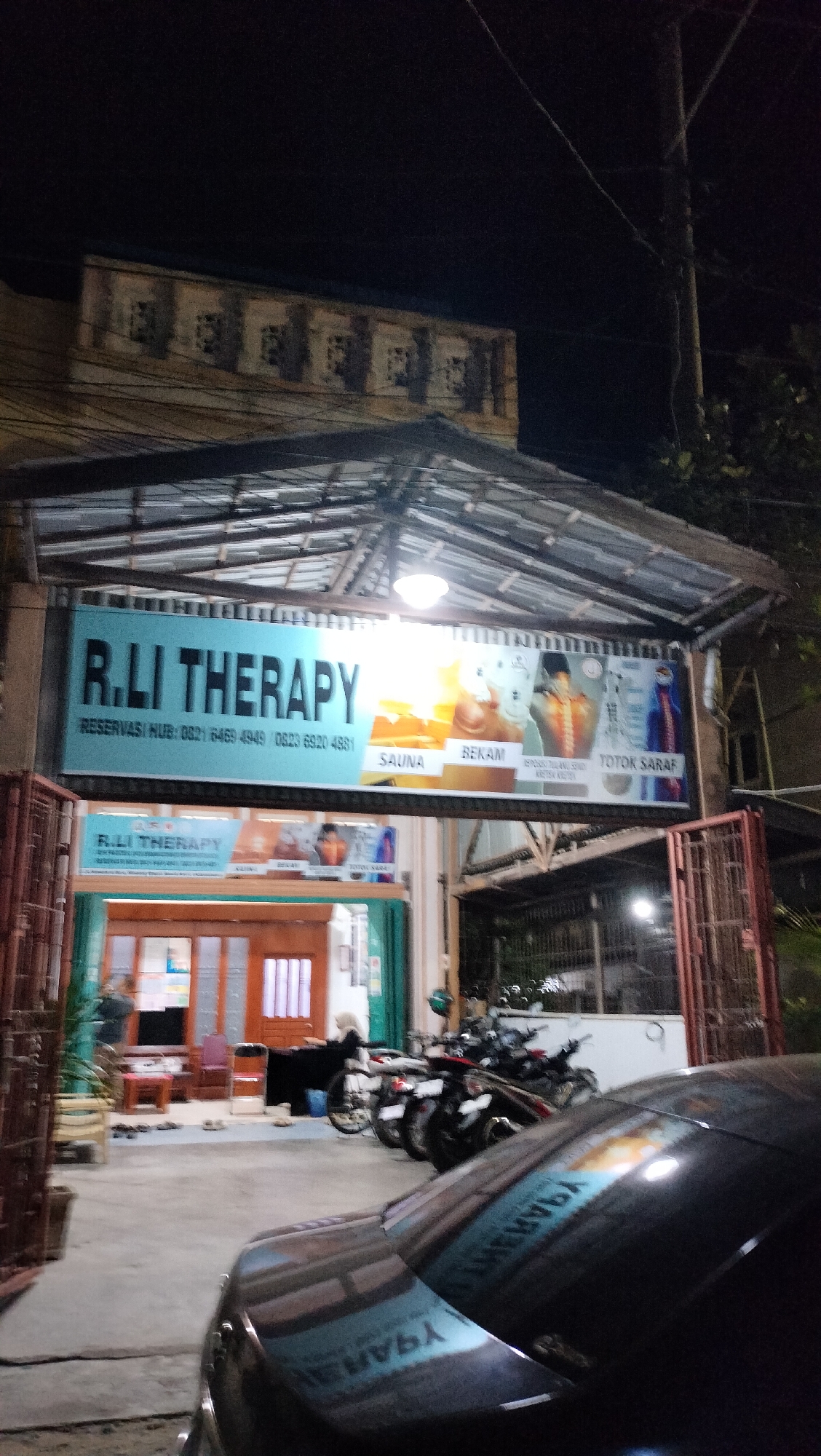 This is a nighttime, eye-level, vertical outdoor shot of a therapy clinic called "R.LI THERAPY" in Lhokseumawe, Indonesia. The clinic is housed in a single-story building with a covered entrance. A prominent, illuminated blue sign hangs above the entrance, displaying the clinic's name in large, bold, black letters. Below the name, in smaller text, it reads "RESERVASI HUB: 0821 6469 4949 0823 6920 4881". The sign also features images illustrating various therapy services, including "SAUNA", "BEKAM", and "TOTOK SARAF".
The entrance itself is framed by red metal gates, one of which is open, revealing a wooden door with glass panels. Inside, the clinic appears to be open, with some interior lighting visible. Several motorcycles are parked outside the entrance, suggesting it's a common mode of transportation for clients. A dark-colored car is partially visible in the foreground, reflecting the clinic's signage. The overall atmosphere is dimly lit, characteristic of nighttime, and there are no discernible weather conditions. The scene appears calm and functional, with no people clearly depicted in an active state within the frame, though a few figures are visible inside the building.