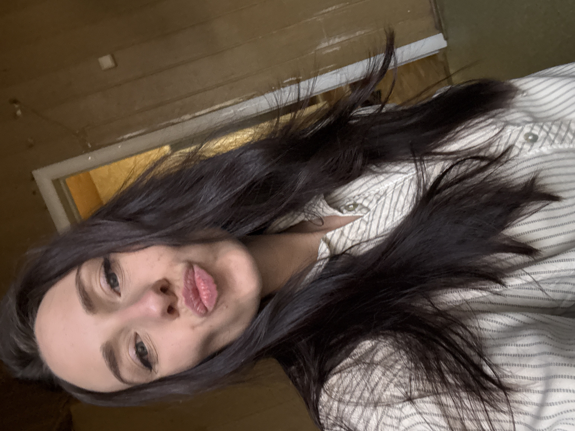 A medium close-up portrait of a young woman with long, dark hair and a light complexion. The image is rotated counter-clockwise, showing her head tilted significantly to the left. She looks directly at the camera with dark eyes, wearing winged eyeliner, and is making a playful "pouty" or "kissing" face with her lips slightly puckered. Her dark hair frames her face and flows over her shoulders, appearing voluminous and somewhat disheveled.
She is wearing a light-colored, possibly off-white or cream, collared shirt with subtle, thin stripes and visible buttons. The background is an indoor setting, characterized by warm brown wooden wall paneling. To her left, a part of a doorway or opening with a lighter-colored frame is visible, leading into a darker, indistinct space. A small, square, light-colored object, possibly a switch, is present on the wooden wall in the upper left background. The lighting appears to be indoor ambient light. The woman's expression conveys a sense of lightheartedness and playfulness. There is no visible text in the image.