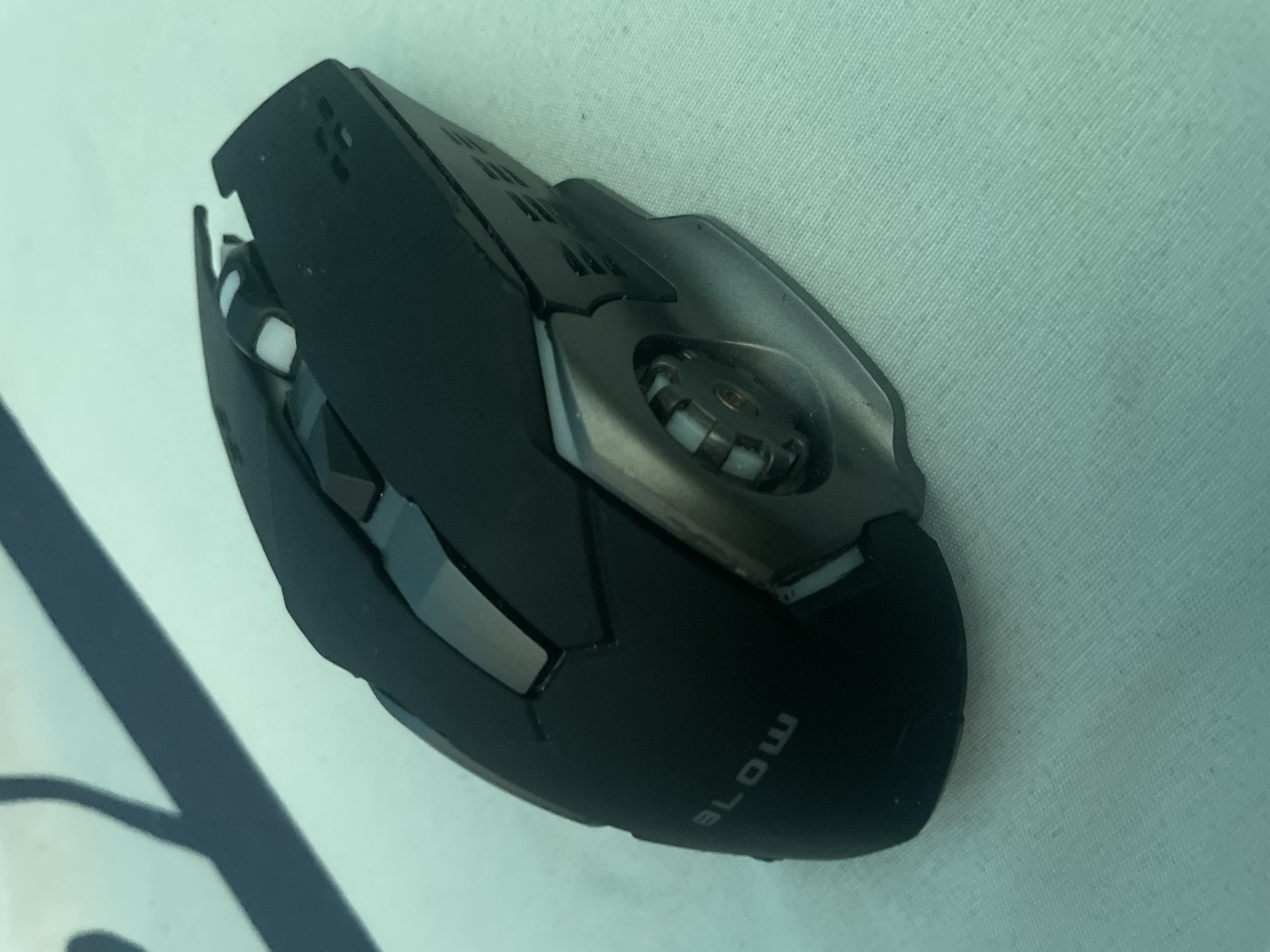 A black gaming mouse with metallic accents is positioned on a light blue textured surface. The mouse has a segmented design, with a prominent scroll wheel and several buttons visible. The word "BLOW" is embossed on the side of the mouse. The background consists of a light blue surface with a subtle striped texture. There are no people present in the image, and the environment is not identifiable as being in Kołobrzeg, Poland. The lighting suggests an indoor setting.