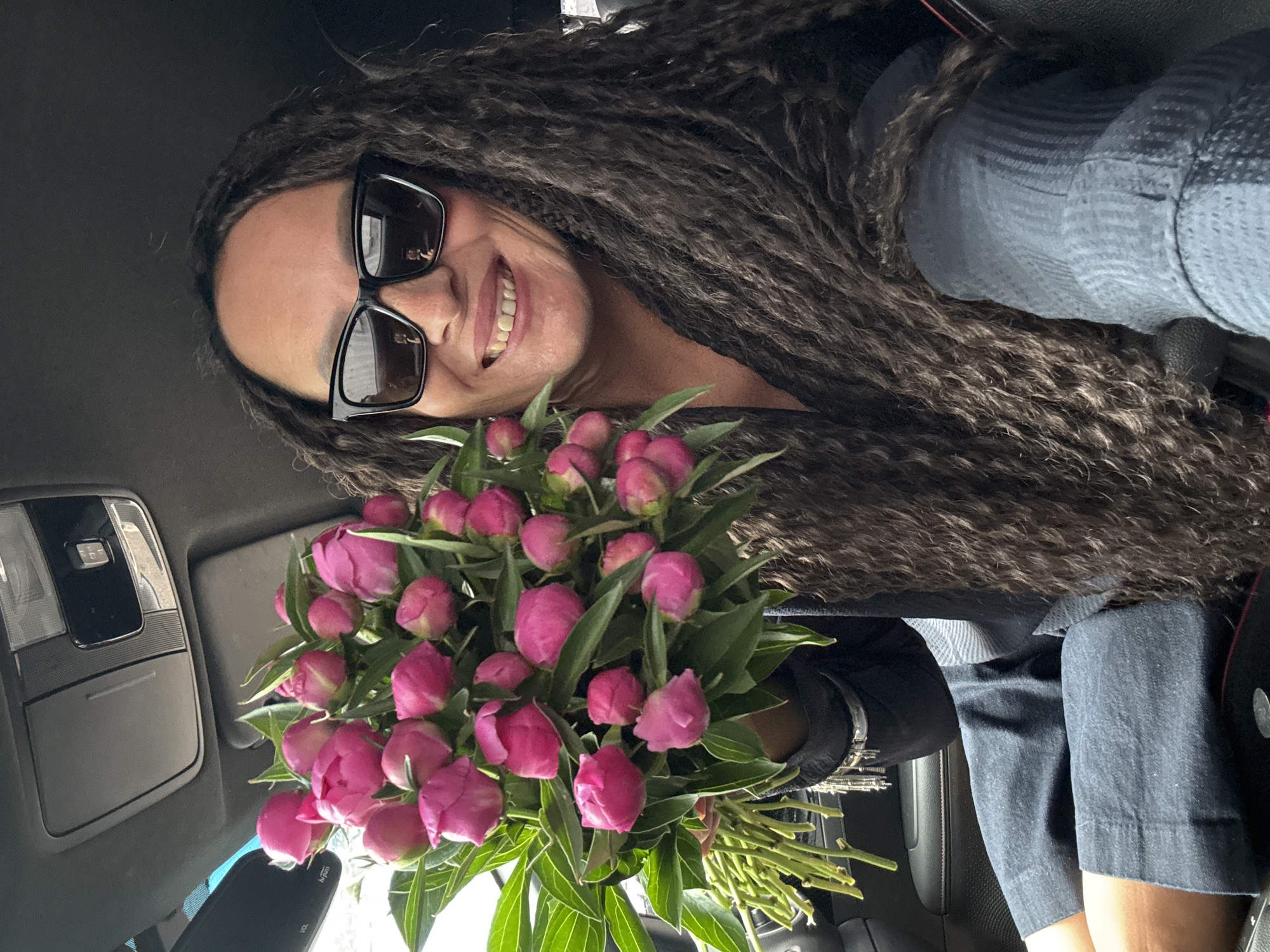 The image captures a smiling woman seated inside a car, holding a generous bouquet of pink peony buds. Her head is tilted, and she looks towards the viewer with a broad, joyful smile, revealing her teeth. She wears dark, rectangular sunglasses that reflect the surroundings. Her long, dark hair, styled in dreadlocks or thick braids, flows over her shoulders and a light blue/grey textured top, worn beneath a darker garment.
The vibrant bouquet, centered in the lower half of the frame, consists of numerous tightly closed pink peony buds, surrounded by fresh green leaves and stems. The setting is the interior of a modern car, with details such as a rearview mirror visible in the upper left, displaying the text "Hi-Pass." An overhead console with lights and a sun visor are also discernible. The bright, natural illumination suggests the scene is taking place during the daytime. The woman's cheerful expression, combined with the fresh flowers, conveys a sense of happiness and celebration. This moment is captured in Chaikovsky, Russia.