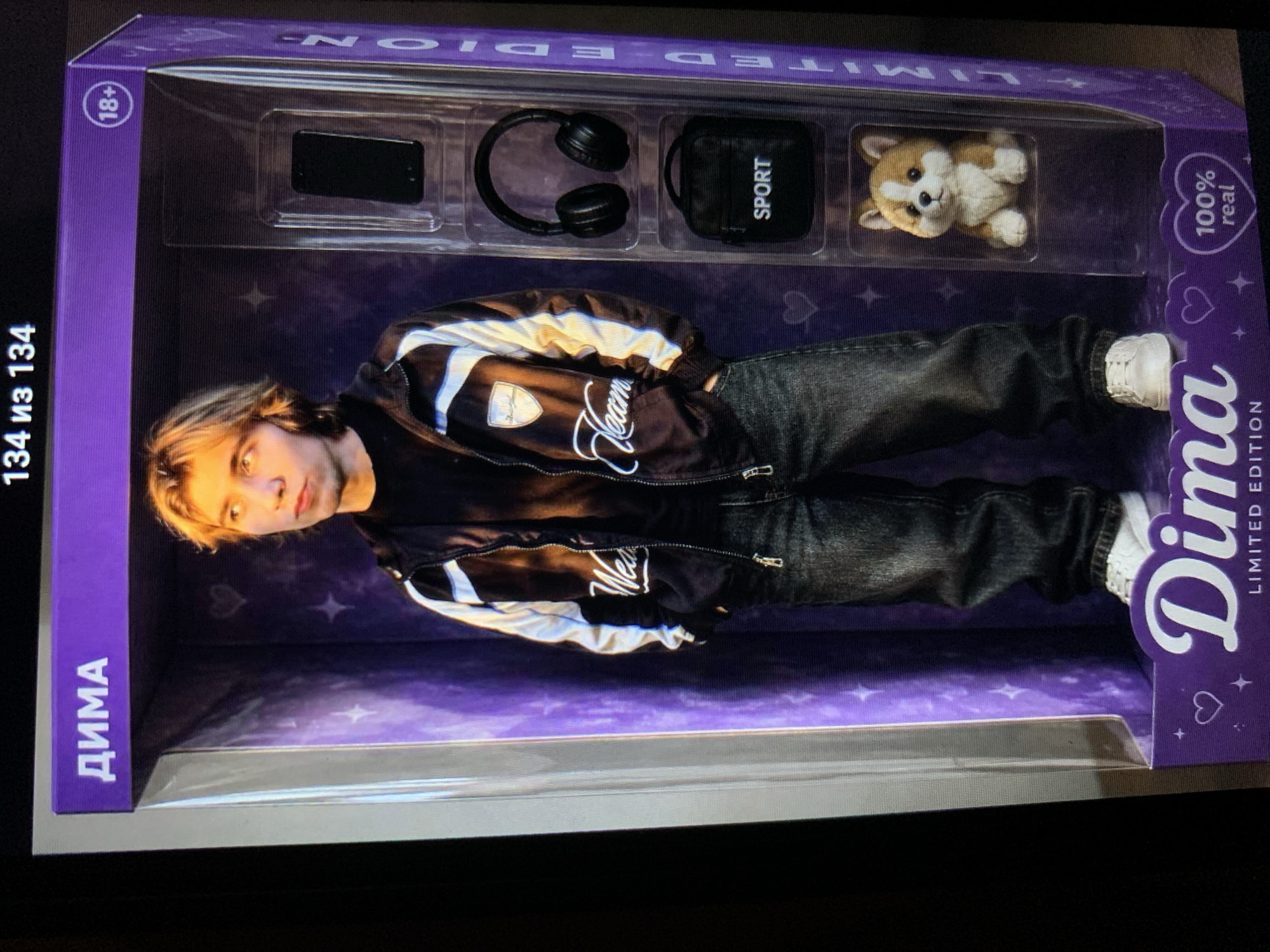 A young man with blond hair, wearing a black and white jacket, a black t-shirt, and dark jeans, stands inside a large, purple doll-like box. The box is designed to resemble a toy package and features graphics such as stars and hearts. On the right side of the box, within separate compartments, are several accessories: a black smartphone, a pair of black headphones, a small black bag labeled "SPORT," and a plush toy dog, possibly a corgi. The man is looking to his right. The text "ДИМА" (Dima) is visible at the top of the box, and "Dima LIMITED EDITION 100% real" is printed on the bottom. The image appears to be a photograph of this display. The context of Lymanka, Ukraine is not discernible from the image itself.