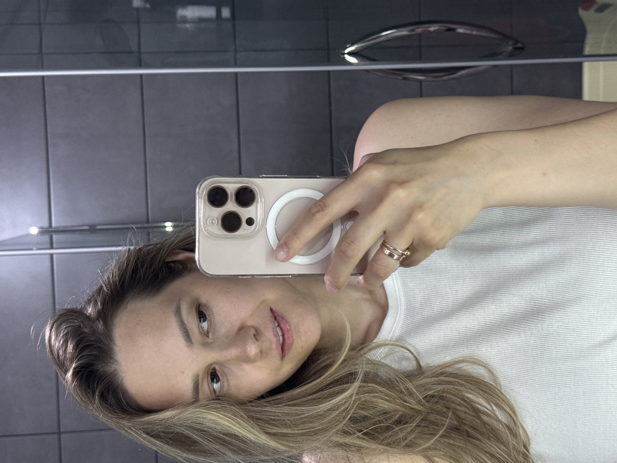 A woman is taking a selfie in a bathroom mirror. The background shows gray tiles and a towel rack. She is holding a phone with a light-colored case and a magnetic ring on the back. She has blonde hair and is wearing a white t-shirt. The location is inferred to be in the city of Chaikovsky, Russia.