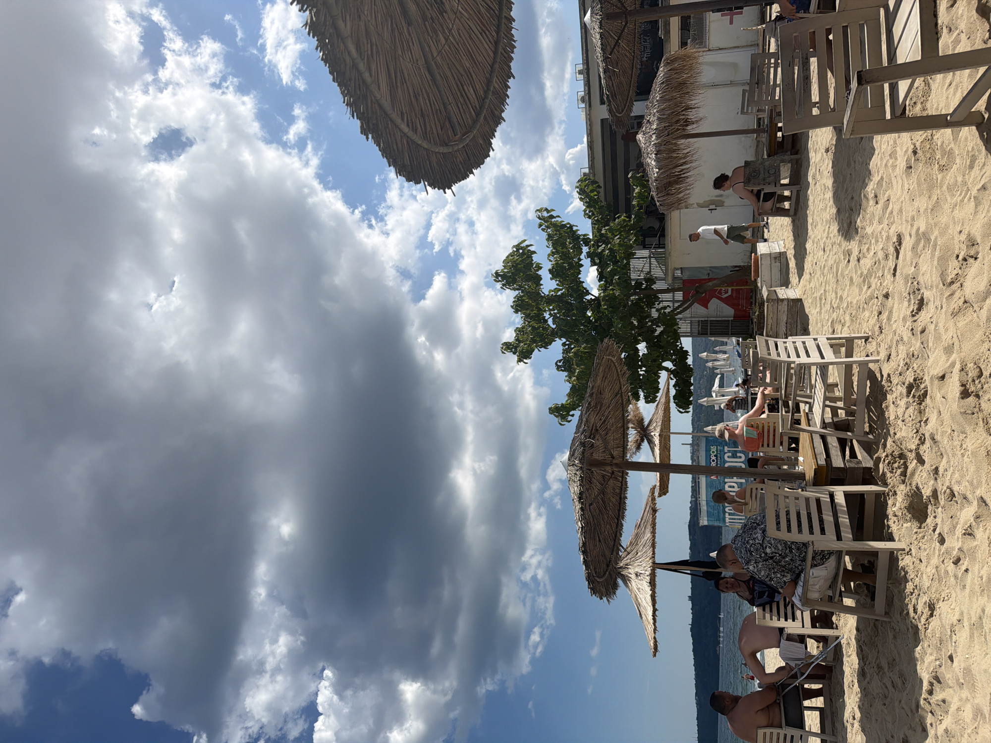 This is an eye-level, medium shot of a beachside cafe in Varna, Bulgaria. The scene is set on a sandy beach with the sea visible in the background. Several thatched umbrellas provide shade over tables and chairs where patrons are seated. Some people are dining, while others are relaxing. A tree with dense green foliage is positioned centrally, with a building and more umbrellas visible behind it. The sky is partly cloudy with a mix of blue sky and dark clouds, suggesting varied weather. In the distance, several boats are visible on the water, and the coastline of Varna is visible across the bay.