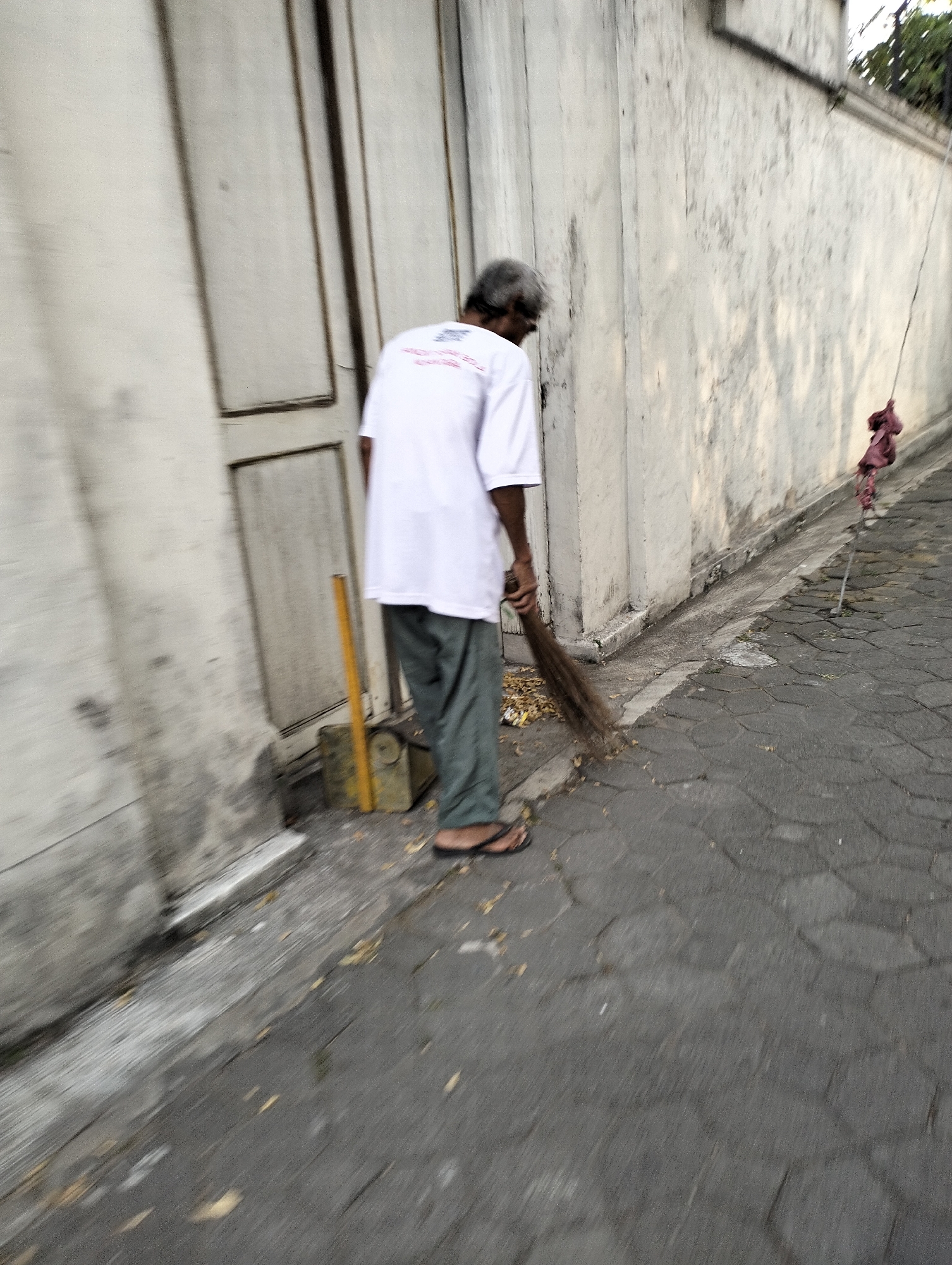 An elderly man with graying hair, wearing a white t-shirt and green pants, sweeps a paved walkway with a broom. He is positioned in the lower half of the frame, facing away from the viewer. To his left, a cream-colored wall with vertical panels forms the background, and a yellow object, possibly a tool or container, leans against it. The walkway is paved with dark, hexagonal stones. Sunlight casts shadows on the wall. Further down the walkway, a thin metal pole with a pink cloth tied to it is visible. The scene appears to be in an outdoor urban environment, possibly in Surakarta, Indonesia, given the architectural style of the wall and the paving stones.