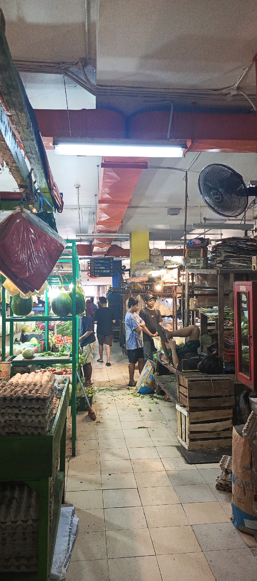 The image depicts an indoor market scene, likely in Sukaresmi, Indonesia, with a focus on fresh produce and general goods. In the foreground, a green stall displays numerous cartons of eggs stacked high. To the left, hanging from a wooden beam, is a large red bag covered in plastic. Further into the market, a display of large green gourds, possibly watermelons or pumpkins, is visible. Beyond this, a variety of red tomatoes and other vegetables are arranged on shelves and in crates.
Several individuals are present in the scene. In the middle ground, three men are engaged in conversation near the produce stalls. One man, wearing a black t-shirt and a cap, stands to the right. To his left, a man in a blue and white tie-dye t-shirt and dark shorts stands, facing the other man. Another man in a black t-shirt and shorts is walking away in the background.
To the right of the produce stalls, a man is lying down on a wooden bench, appearing to be resting or looking at his phone. He is wearing dark shorts and a dark t-shirt. In front of him, several wooden crates are stacked. Behind him, there are shelves laden with various items, including what appear to be smaller containers and possibly woven baskets. A large industrial fan is mounted on the wall in the upper right portion of the image.
The market floor is tiled with light-colored square tiles. Debris, appearing to be vegetable trimmings, is scattered across the floor in several areas. The ceiling features exposed beams and conduit, with fluorescent lighting illuminating the space. Signs with Indonesian text are visible in the background, indicating different sections of the market, such as "Food Court," "Fashion," and "Toilet." The overall atmosphere suggests a bustling, functional marketplace.