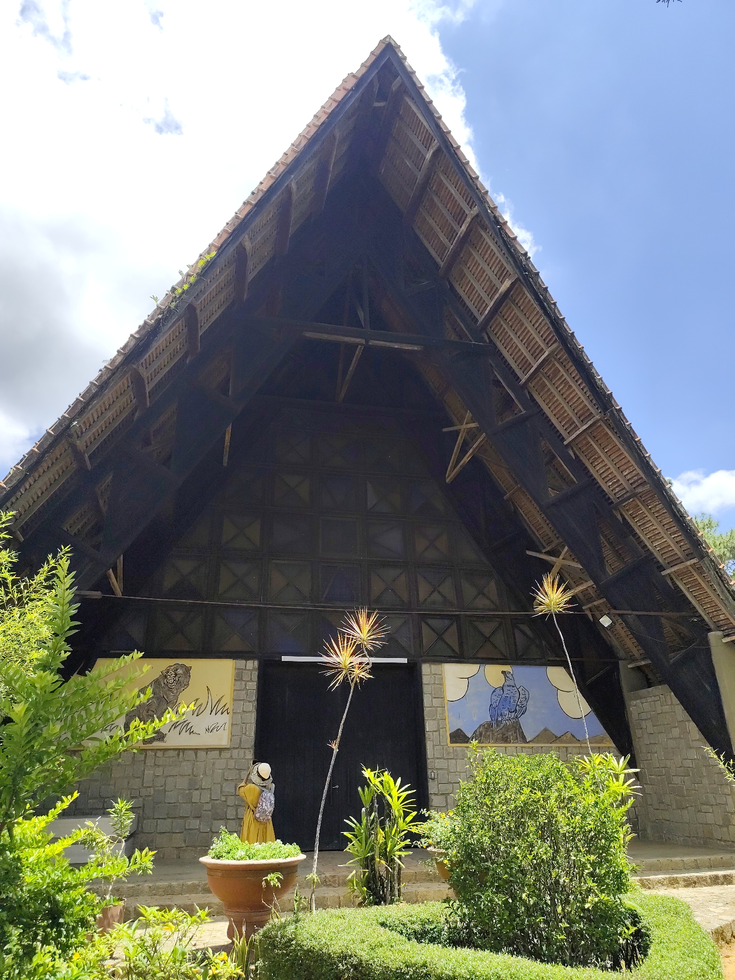 This image depicts the exterior of a building with a steeply pitched, thatched roof supported by exposed wooden beams. The front of the building features a large black door flanked by two murals on a stone facade. The mural on the left depicts a roaring tiger, while the mural on the right shows a blue bird perched on a rock against a backdrop of clouds and mountains. In front of the building, a statue of a person in a yellow robe stands near a large terra cotta planter filled with greenery. Lush vegetation, including bushes and trees, surrounds the building and the statue. The scene is set against a bright blue sky with scattered white clouds, suggesting a sunny day. The architectural style and natural setting are characteristic of the Da Lat region in Vietnam, known for its French colonial influences and lush highlands.