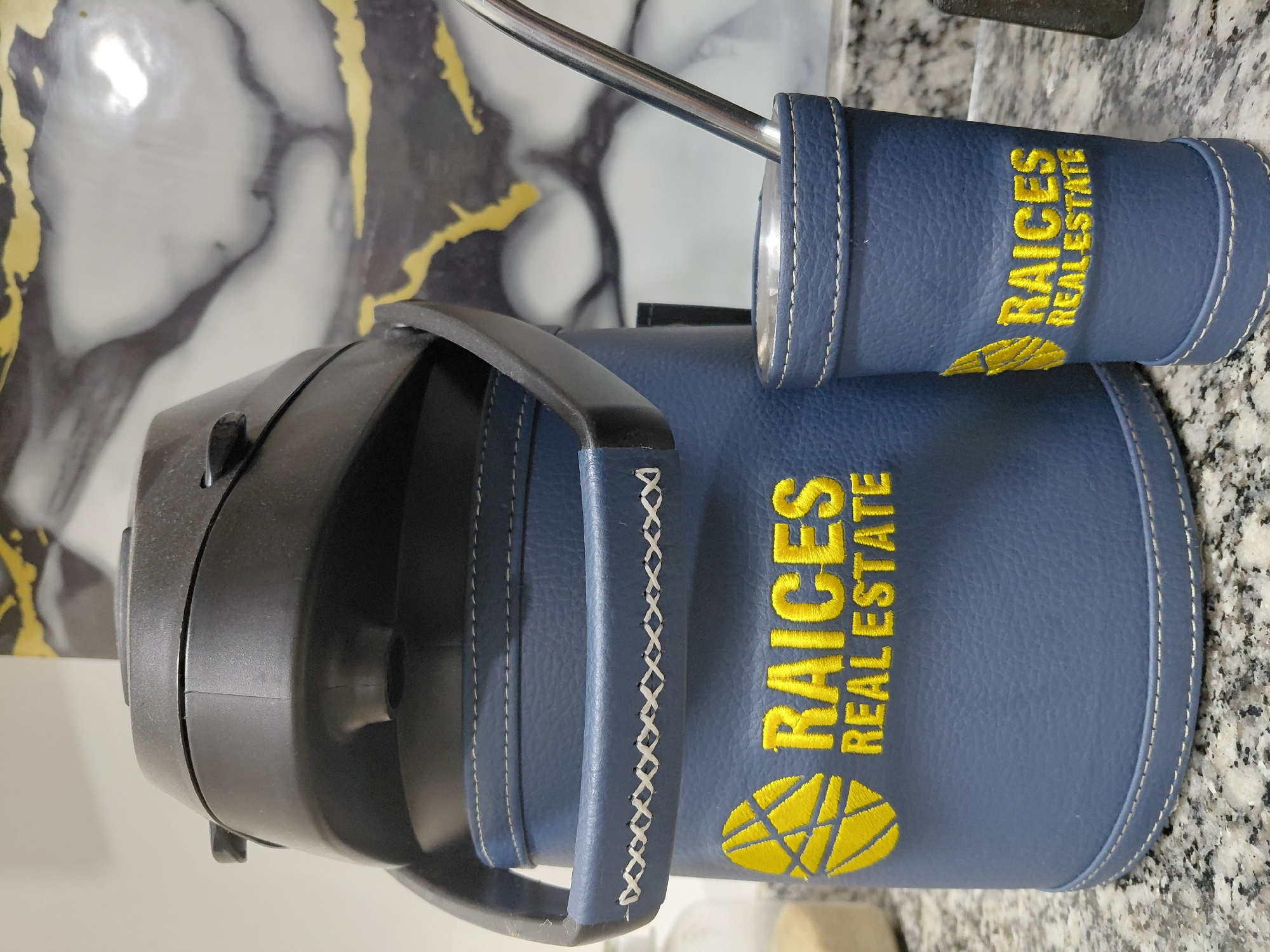 The image shows a close-up of a blue leather-covered thermos and a matching cup with a metal straw, possibly for drinking mate. Both the thermos and the cup are embroidered with a yellow logo and the words "RAICES REAL ESTATE". The background features a marbled surface with gold accents, suggestive of a modern interior design. The objects are placed on a speckled granite countertop, typical of kitchens or commercial spaces. The lighting is neutral, and there are no people visible in the frame. The setting could be a home or office in Encarnación, Paraguay, a city known for its scenic waterfront and growing real estate market, where businesses like "Raices Real Estate" would be relevant.
