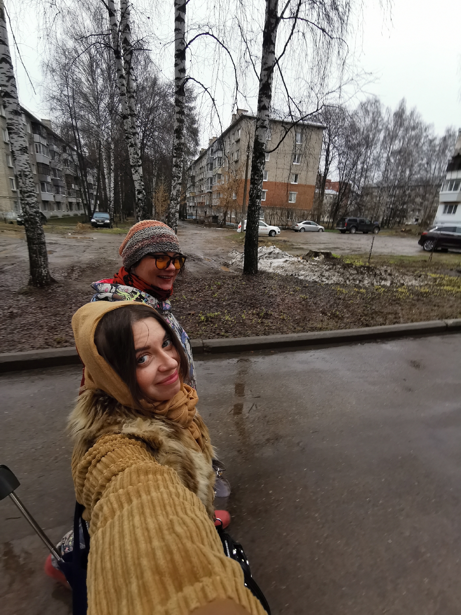 This media file presents a candid, selfie-style photograph of two women walking in an urban residential area of Chaikovsky, Russia. The foreground woman, with dark hair framed by a mustard yellow headscarf and wearing a similarly colored ribbed coat with fur trim, smiles directly at the camera. Behind her, another woman, wearing a colorful knitted beanie and yellow-tinted sunglasses, also smiles as she looks towards the right. A part of a stroller handle is visible on the far left, suggesting a family outing.
The scene features several multi-story apartment buildings, typical of post-Soviet architecture, with many windows. The area is populated by numerous birch trees with bare branches, indicating a cold season such as late autumn, winter, or early spring. The ground consists of wet asphalt and muddy patches with sparse, dead leaves and remnants of snow or slush, consistent with recent precipitation or melting. The overcast sky provides soft, diffused light, suggesting it is daytime.
Several parked cars are visible in the background, including a white vehicle on which the partial text "4.99.99" can be seen, possibly indicating a service number. Both women appear cheerful, conveying a positive mood despite the somewhat grey and damp weather conditions. The image captures a casual moment in a characteristic Russian neighborhood.