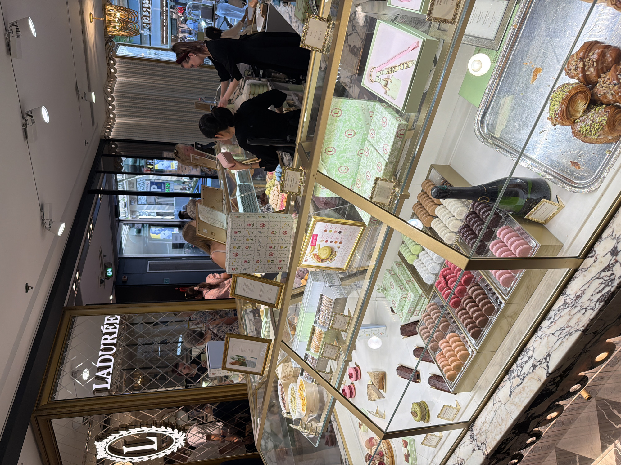 The image shows the interior of a Ladurée shop, likely located in Paris, France, based on the brand's origin and prominence there. A display case filled with pastries, cakes, and macarons dominates the foreground. The display case has a gold frame. Boxes of pastel macarons are visible, stacked neatly in the case. A bottle of Champagne is in the display case. Croissants are displayed on a silver tray. People, including staff and customers, are visible behind the counter, some wearing dark clothing. The shop has an upscale, elegant aesthetic, with decorative lighting, marble countertops, and mirrored surfaces that reflect the exterior. A sign with "LADURÉE" is prominently displayed near the entrance.