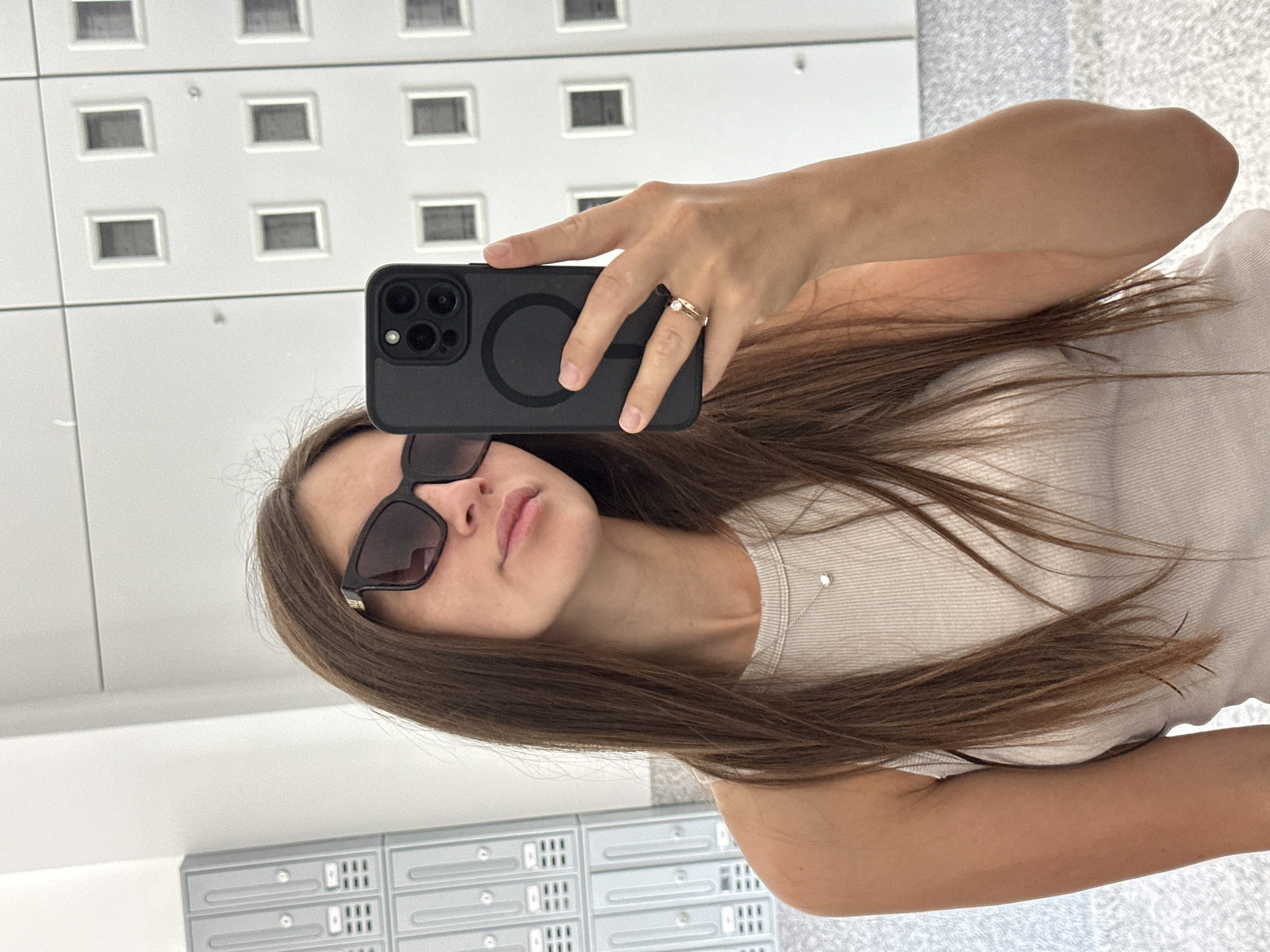 A young woman is taking a selfie in a modern hallway with a tiled wall and numerous small, rectangular windows, possibly mailboxes or ventilation shafts. She is wearing a light-colored tank top and large, dark-rimmed sunglasses. A delicate silver necklace with a small pendant rests on her collarbone. Her long, brown hair is down and cascades over her shoulder. In her right hand, she holds a black smartphone, the camera facing her. A simple gold ring with a small diamond is visible on her left ring finger. The lighting appears bright and even, suggesting it's daytime. The environment seems clean and well-maintained. The overall tone is casual and self-aware.