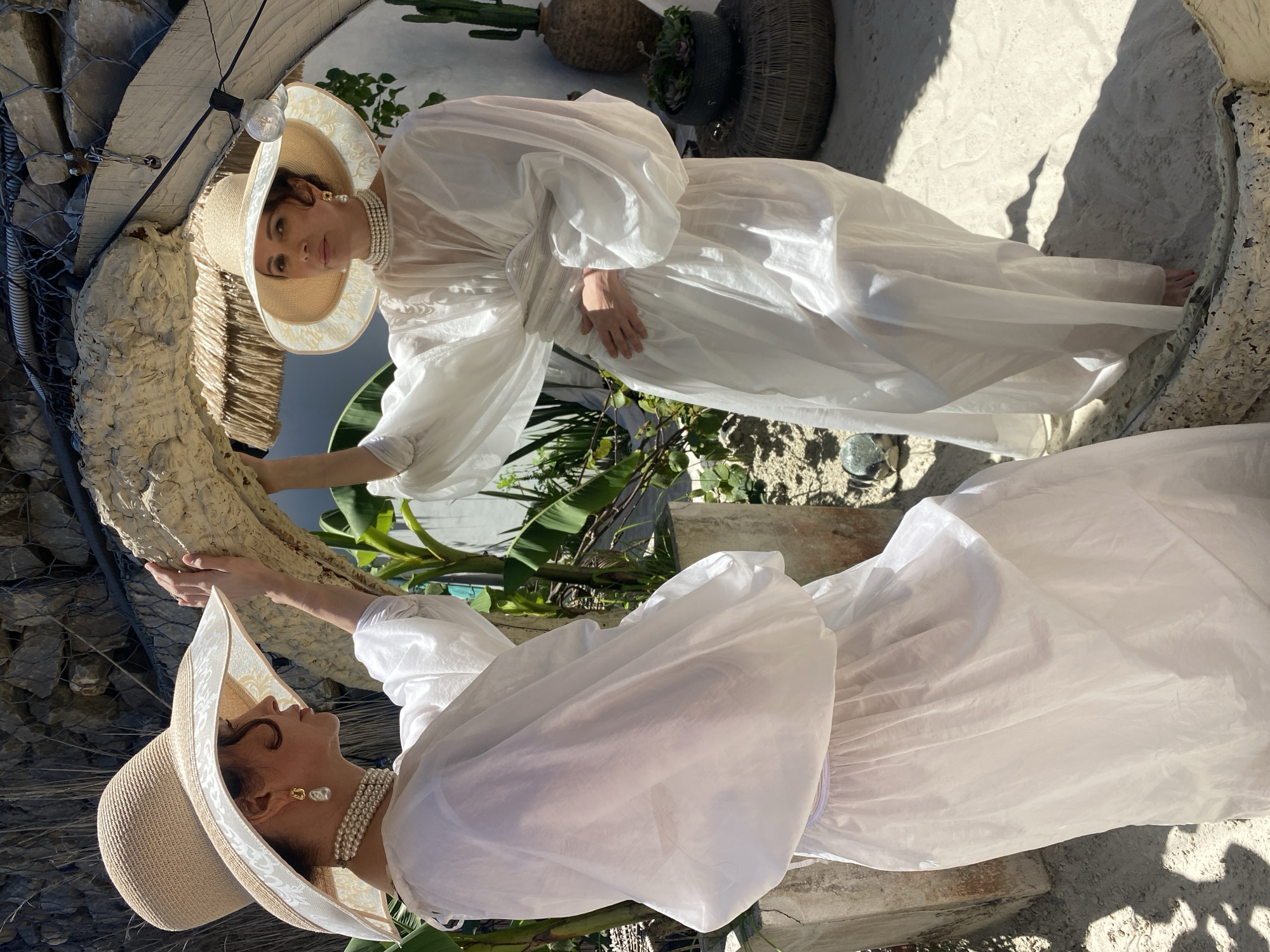 Two women are standing outdoors in what appears to be a sun-drenched, sandy area with lush greenery and a stone wall in the background. The woman on the left is facing away from the camera, leaning her left hand against a roughly textured, large, light-colored object, possibly a sculpture or natural formation. She is wearing a wide-brimmed straw hat with a decorative band, a white flowing dress with voluminous sleeves, and pearl jewelry, including a choker necklace and large earrings. The woman on the right is facing the camera, her right hand resting on her abdomen. She is also wearing a similar wide-brimmed straw hat, a white flowing dress, and pearl jewelry. The environment includes plants, a rustic thatched roof structure, and a light bulb hanging from a wire overhead. The ground is covered in sand, with shadows cast by the sun. The scene suggests a relaxed, possibly resort-like setting. The location is not specified within the image itself, and it is not possible to determine if it is in Sochi, Russia.