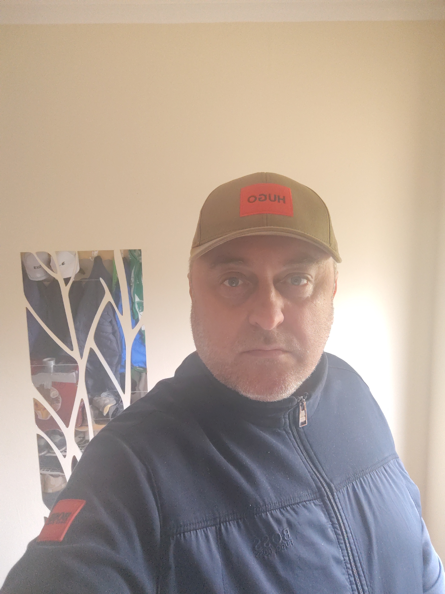 A middle-aged man is captured in a head-and-shoulders indoor selfie, looking directly at the camera with a neutral, serious expression. He has short, receding grey hair and light stubble on his face. He is wearing a dark olive green or khaki baseball cap featuring a red rectangular patch on the front. The patch displays the word "HUGO" in white capital letters, which appears mirrored as "OGUH," suggesting a front-facing camera.
The man is dressed in a dark blue or navy zipped jacket with visible stitching and what appears to be a quilted or padded upper section. A second red rectangular patch, also with the mirrored "HUGO" text, is visible on his left sleeve. Further mirrored text, likely "BOSS" and potentially "HUGO BOSS," is subtly visible on the chest area of his jacket.
Behind him, to the left, is a tall, narrow, decorative object, possibly a mirror or wall art, featuring white, tree-branch-like cutouts against a reflective surface. Through the reflective gaps, various items of clothing or accessories on a rack are vaguely visible, including white and blue garments. The background consists of a plain, light-colored wall, likely a warm off-white or pale beige. The scene is lit by general indoor lighting. The image provides no specific time of day or weather cues, but the context indicates the location is Huntingdonshire, United Kingdom.