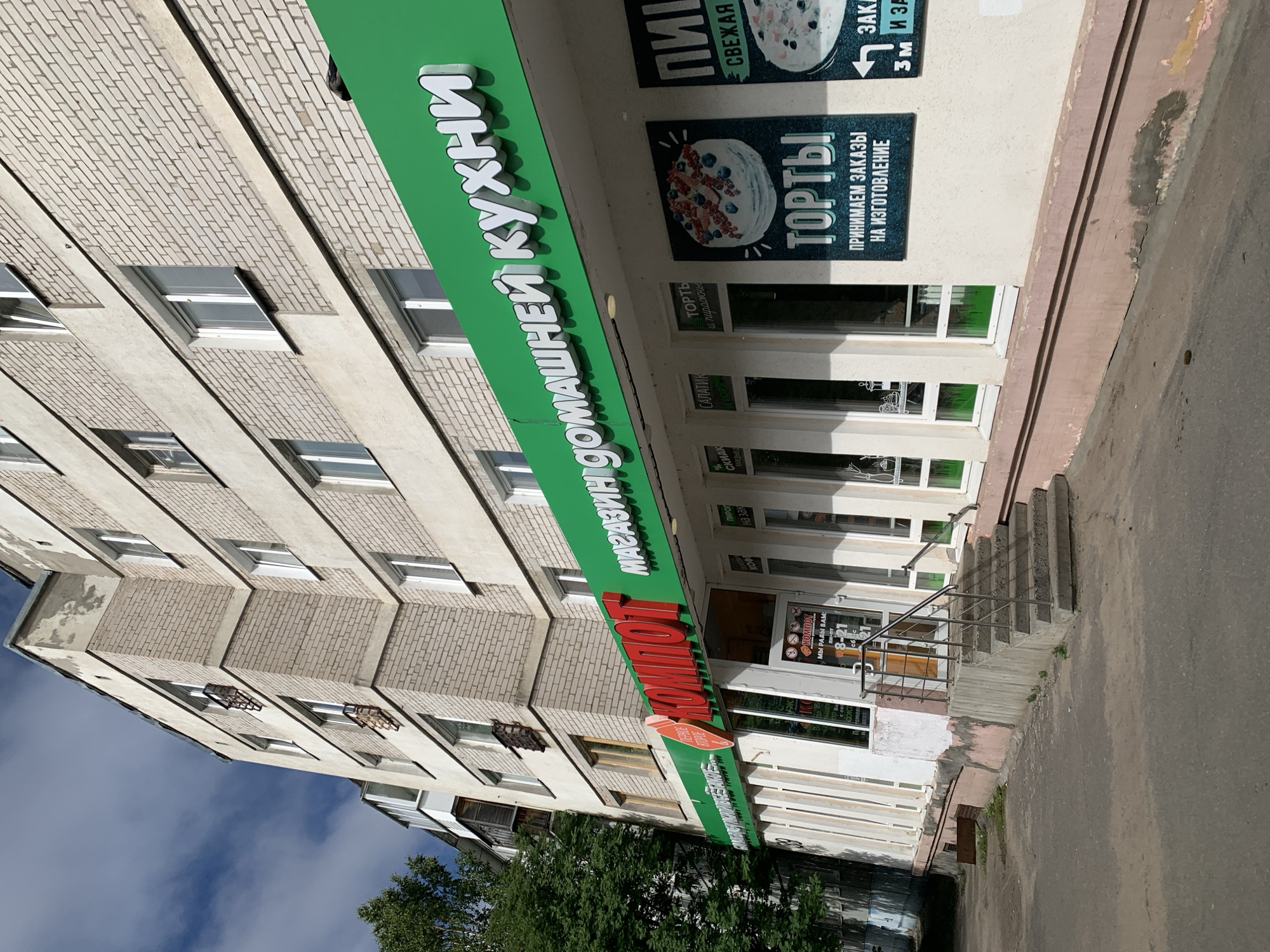 This media file is a photograph taken from a low angle, looking up at the facade of a multi-story building. The building appears to be an apartment building or a commercial establishment with storefronts on the ground floor. The facade is constructed of light-colored brick with horizontal bands of concrete. Several windows are visible, some with closed blinds and others with open sections.
A prominent green sign with white, three-dimensional lettering stretches across the upper portion of the building, displaying the text "МАГАЗИН ДОМАШНЕЙ КУХНИ" (translated as "Homemade Food Store"). Below this, another sign with red lettering reads "КОМБОТ" (KOMBOT). Adjacent to the main entrance, there's a smaller sign with business hours: "МЫ РАДЫ ВАМ: пн-пт 8-21" (We are happy to see you: Mon-Fri 8-21).
Several smaller signs with images of food items, such as cakes and salads, are also visible near the entrance, indicating the nature of the business. The building is situated on a paved surface, likely a sidewalk or street, with a concrete staircase leading up to the entrance. Trees and vegetation are visible on the right side of the frame, suggesting an outdoor setting. The sky is visible in the upper left corner, appearing partly cloudy and blue, indicating daytime. The lighting suggests it is a sunny day. The overall scene depicts the exterior of a commercial establishment in a residential or mixed-use area.