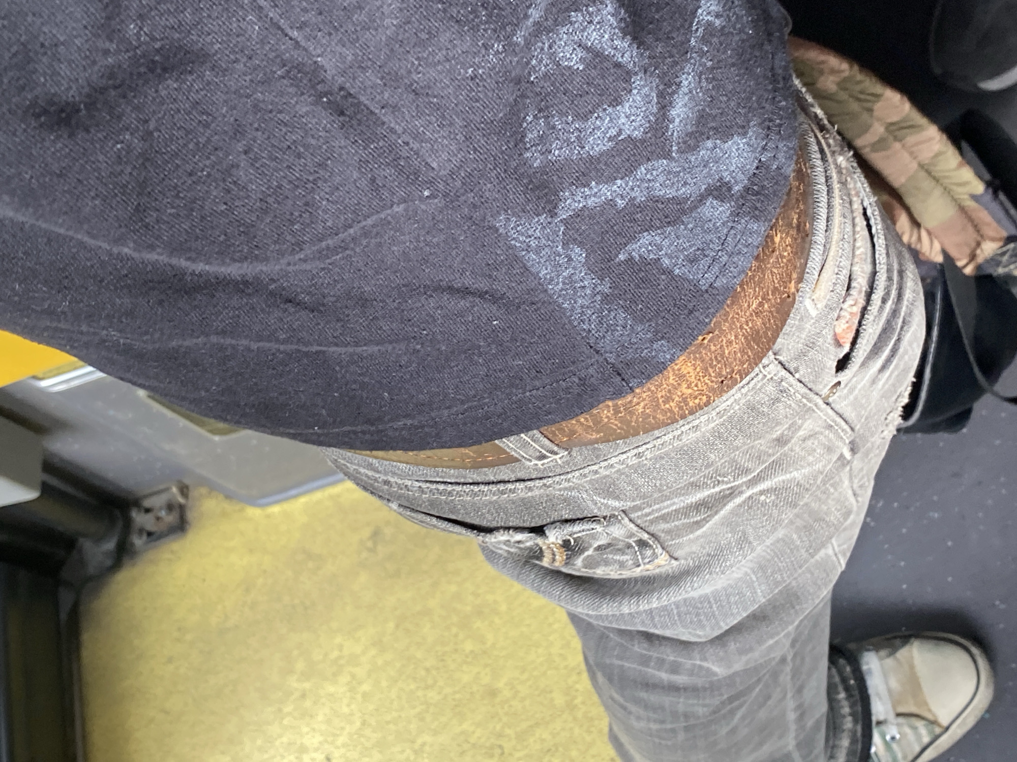 A close-up, overhead shot shows a person's legs and torso, seated on what appears to be public transportation. The person is wearing faded grey jeans, a dark t-shirt with a distressed graphic, and a brown belt. The t-shirt has a light-colored, faded design on the right side. One foot, clad in a light-colored sneaker with green and beige accents, is visible on the left. The background consists of a yellow-colored floor, likely the interior of a bus or train, with metal fixtures and dark flooring. The scene is devoid of any specific identifiable landmarks or characteristics that would pinpoint its location within Almaty, Kazakhstan.