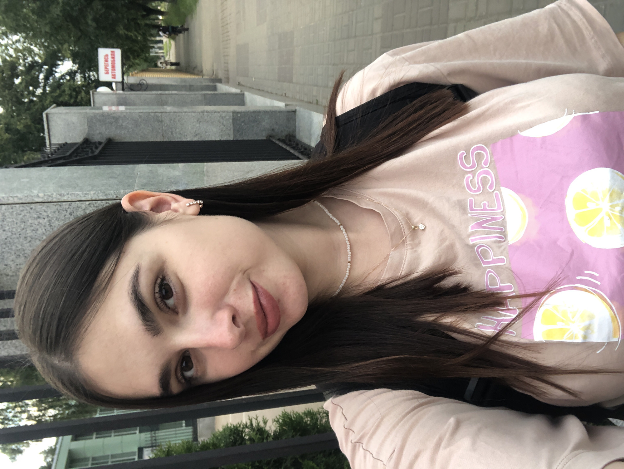 A young woman with long, dark hair is taking a selfie outdoors. She is wearing a light pink t-shirt with a graphic print of lemons and the word "HAPPINESS" in pink lettering. She has on a delicate pearl necklace with a small pendant and small hoop earrings. She is standing on a paved sidewalk next to a black metal fence and a stone wall. In the background, a building with green accents and trees are visible. A sign in Russian that reads "БЕРЕГИТЕСЬ АВТОМОБИЛЕЙ" (Beware of Cars) is mounted on the wall. Further down the sidewalk, a person is walking and another person is riding a bicycle. The scene appears to be in the city of Chaikovsky, Russia, suggested by the Russian signage and typical urban architecture.