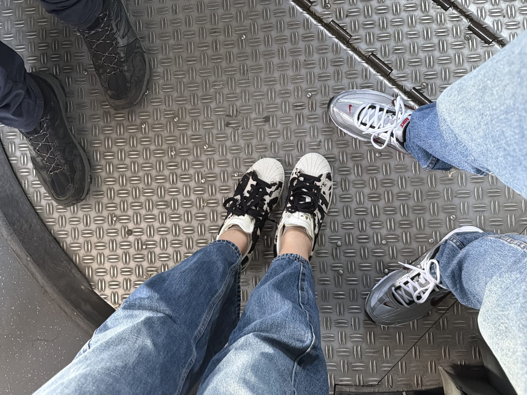 The image shows an overhead view of four pairs of legs and shoes standing on a metal platform. The legs are wearing jeans. The shoes are different styles: dark hiking boots, Adidas Superstar with a black and white pattern, silver Nike sneakers, and gray sneakers. The metal platform has a textured surface. The setting appears to be an industrial or public transport environment. Without additional information, it is impossible to confirm the location as Sofia, Bulgaria.