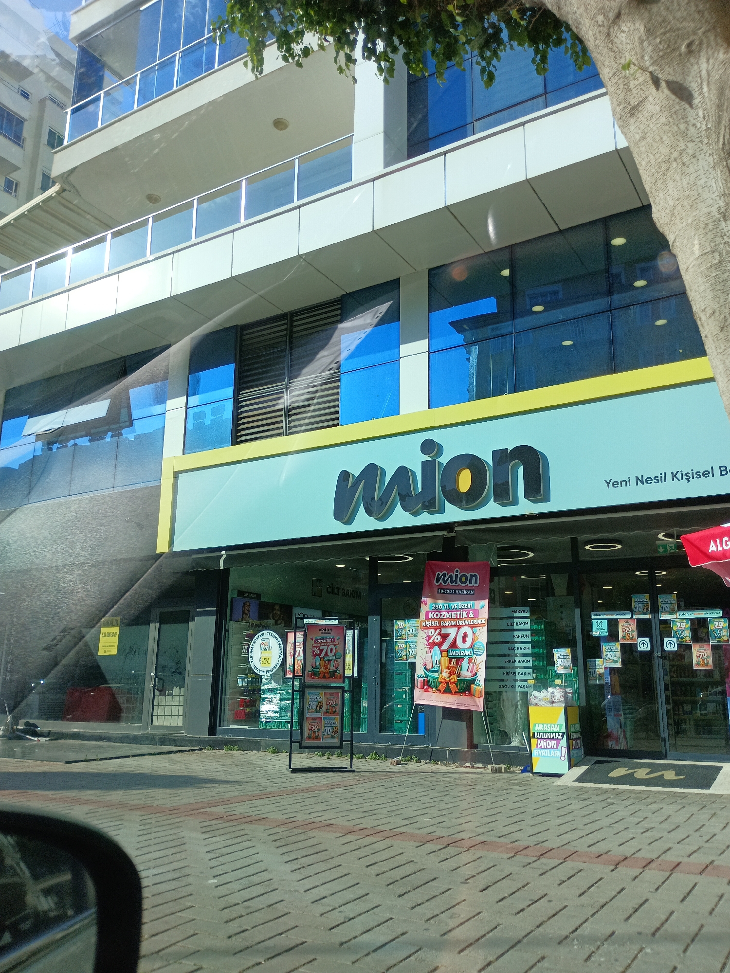 This is a medium-angle, eye-level, exterior shot of a "mion" store, a cosmetics and personal care retailer, on a sunny day in Alanya, Turkey. The store's facade is modern, with large blue glass windows reflecting the sky and surrounding buildings. A prominent light blue sign with the "mion" logo in dark, stylized lettering stretches across the storefront, with the tagline "Yeni Nesil Kişisel Be" (New Generation Personal Be) to its right.
In front of the store, a large promotional poster advertises a "70% discount" on cosmetics and personal care products for purchases over 250 TL, valid from June 19-21. Another display promotes the "mion" brand, stating "What you're looking for, you won't find at mion at these prices!"
Several other smaller signs with "70%" markings are visible on the storefront windows, indicating ongoing sales. The store entrance is partially visible, with a black doormat featuring the "mion" logo. The sidewalk in front of the store is paved with grey bricks. A portion of a large tree trunk is visible on the right side of the frame, its leaves casting shadows on the building. The reflection of a car's side mirror and dashboard is visible in the lower left corner of the image, suggesting the photo was taken from inside a vehicle. The time of day appears to be afternoon, given the sunlight and shadows.