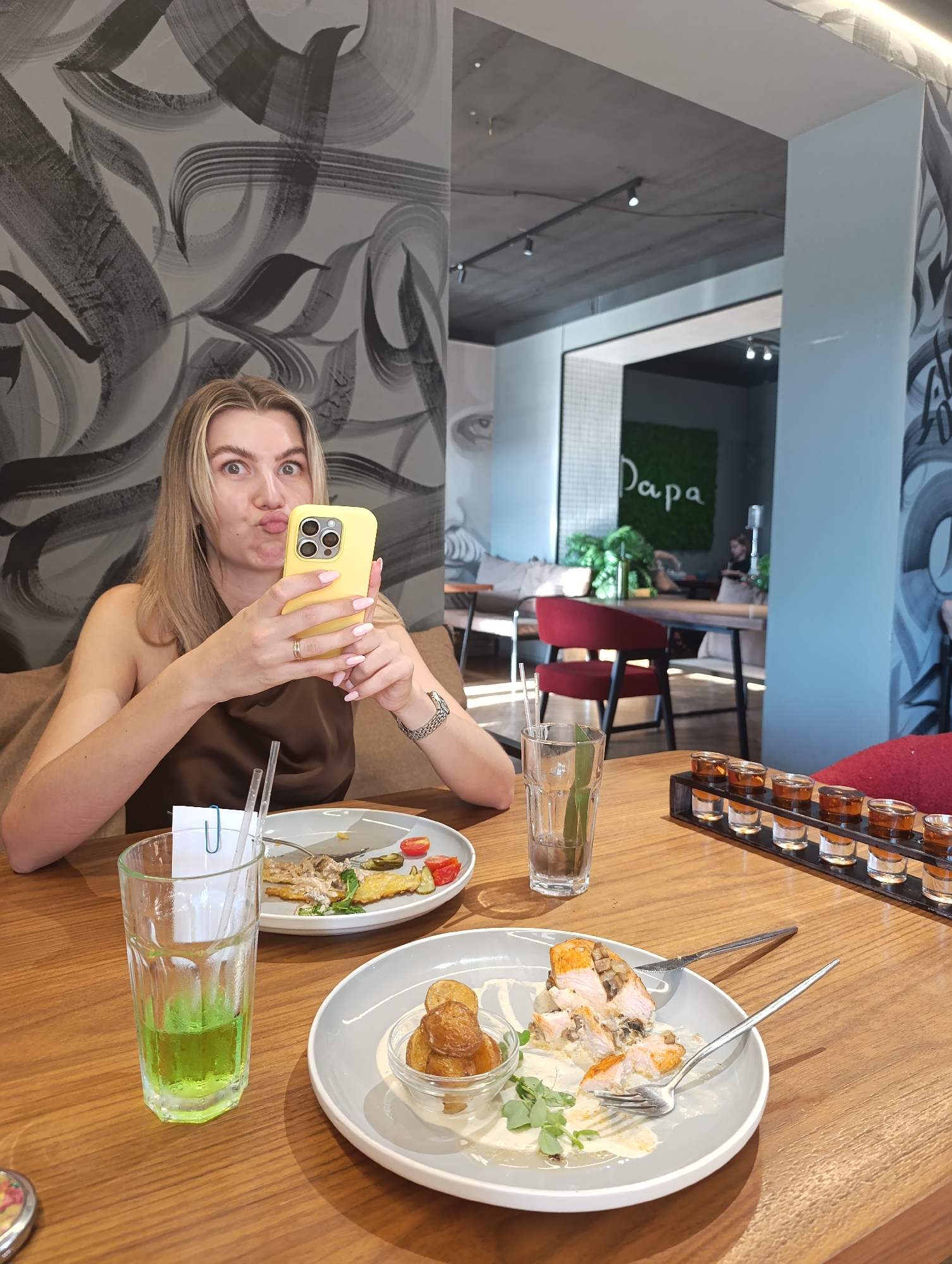 The image depicts a woman sitting at a wooden table in what appears to be a restaurant. She is holding a yellow smartphone in her hands and making a playful expression, as if taking a selfie. In front of her are two plates with food remnants, a glass with a green beverage, and a shot glass rack with several shots. The background shows a wall with a distinctive abstract mural and a glimpse of the restaurant's interior, including red chairs and plants. A sign that reads "Papa" is visible on a green wall in the distance. The lighting suggests an indoor environment during daylight hours.