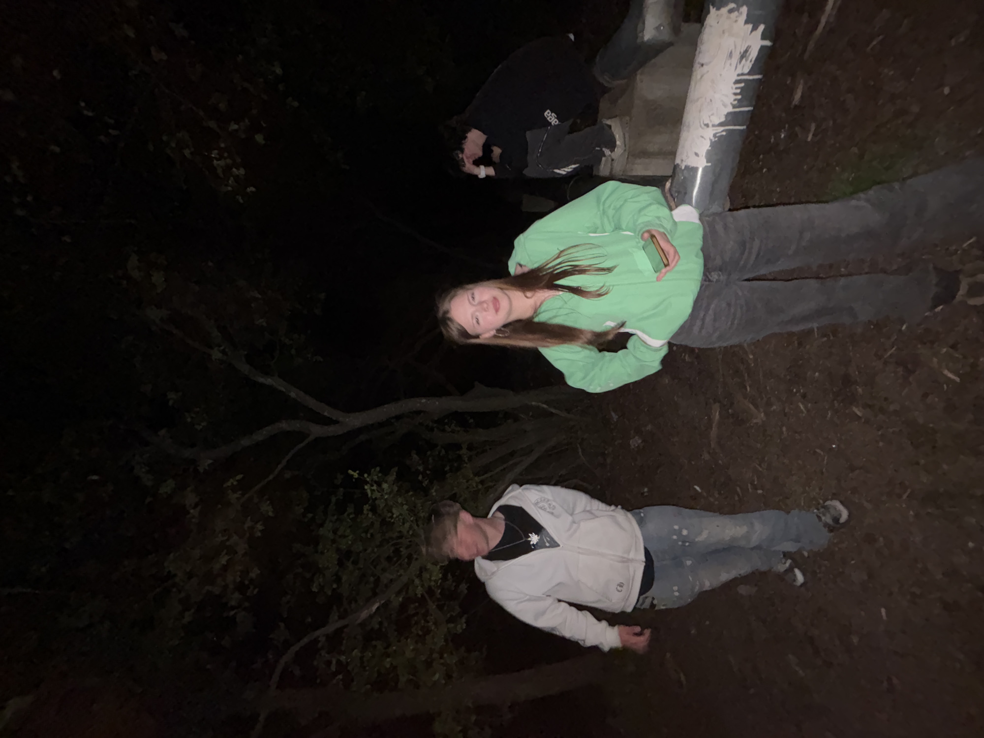 The image depicts a dark outdoor scene in what appears to be a wooded area. Three individuals are present. One person is kneeling on what looks like a concrete structure or base, partially obscured in the darkness. A second individual, wearing a light green sweatshirt, is lying on the ground near the concrete base. A third person, wearing a light-colored jacket, is lying on the ground at the bottom of the frame. The location is unspecified, and there's no evidence to suggest the location is in the city of Фоки, Russia. More data needed to provide a useful response.