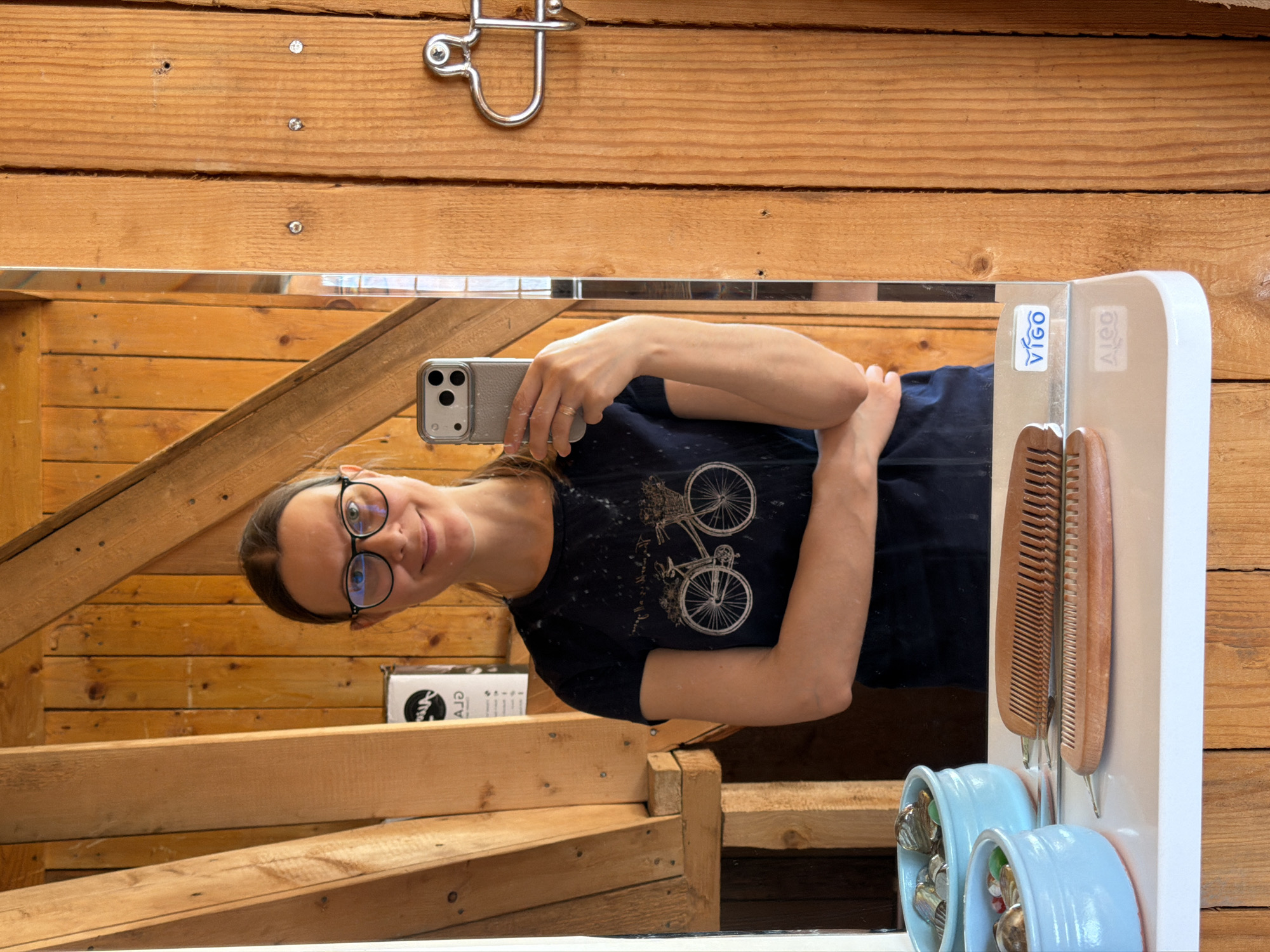 The image shows a selfie taken in a bathroom. The background consists of natural wood paneling. A woman is visible in the mirror's reflection, holding a smartphone. She wears glasses and a dark-colored t-shirt with a bicycle design. Beside the mirror is a white fixture with wooden combs hanging on it. The lower part of the fixture contains two light blue holders, each filled with jewelry. The bathroom's environment is suggested by the wood paneling and bathroom accessories. There is no verifiable indication that the image was taken in the city of Ягул, Russia.