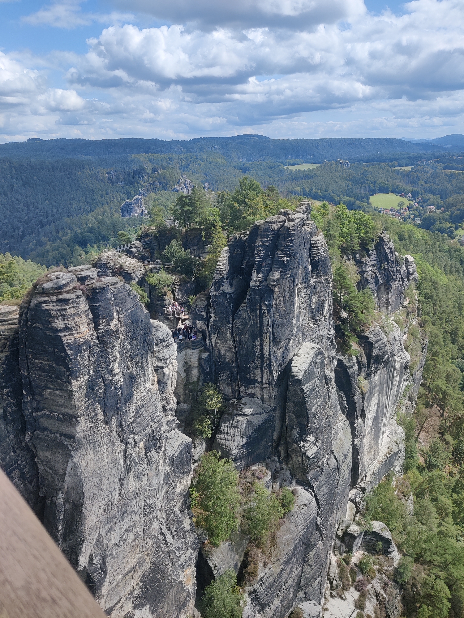 In Lohmen, Germany, the image captures a scenic view of the Bastei rock formation, a prominent sandstone feature in Saxon Switzerland National Park. Jagged, weathered rock spires dominate the foreground, casting shadows and showcasing intricate geological layers. A wooden railing is visible in the lower left corner, suggesting a viewpoint. On a bridge connecting some of the rock formations, a group of approximately twenty people is gathered, observing the landscape. They appear to be tourists, dressed casually in clothing suitable for outdoor activities. The background reveals a vast expanse of dense green forest, interspersed with rolling hills and a distant village with red-tiled roofs. The sky above is a mix of blue and white clouds, indicating daytime and fair weather. The overall scene conveys a sense of natural grandeur and recreational tourism.