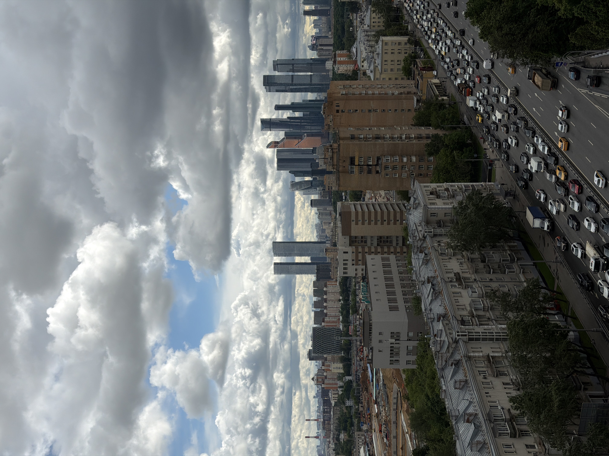 Here's a description of the image:
The image shows a high-angle, somewhat rotated view of a cityscape under a cloudy sky. The sky dominates the left portion of the image, filled with grey and white clouds, with patches of blue sky visible.
In the center of the image, a cluster of tall buildings is identifiable as the Moscow International Business Center, often called Moscow City. Lower residential and commercial buildings are positioned closer to the viewer.
To the right of the buildings, a major road is visible, with heavy traffic congestion. Cars and trucks are lined up in multiple lanes, indicating a busy transportation artery. Buildings and trees line the road.
In the foreground, construction or excavation is occurring. Equipment is visible, indicating ongoing work.