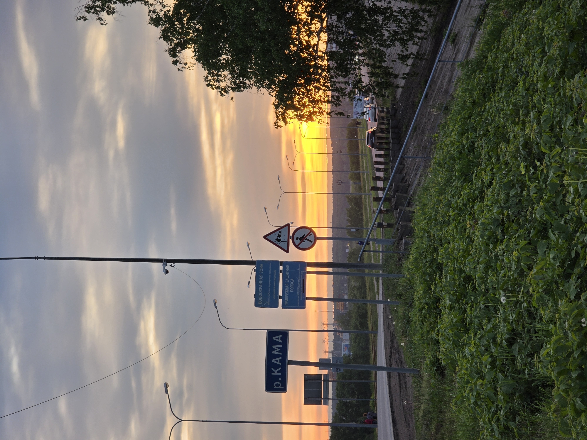 The image shows a sunset in Chaikovsky, Russia. The sky is filled with clouds and orange light. In the foreground, there is lush greenery. There are also several lampposts and road signs, including a sign that reads "р. КАМА" (r. KAMA), which likely refers to the Kama River. Other signs are in Russian. A section of road is also visible.