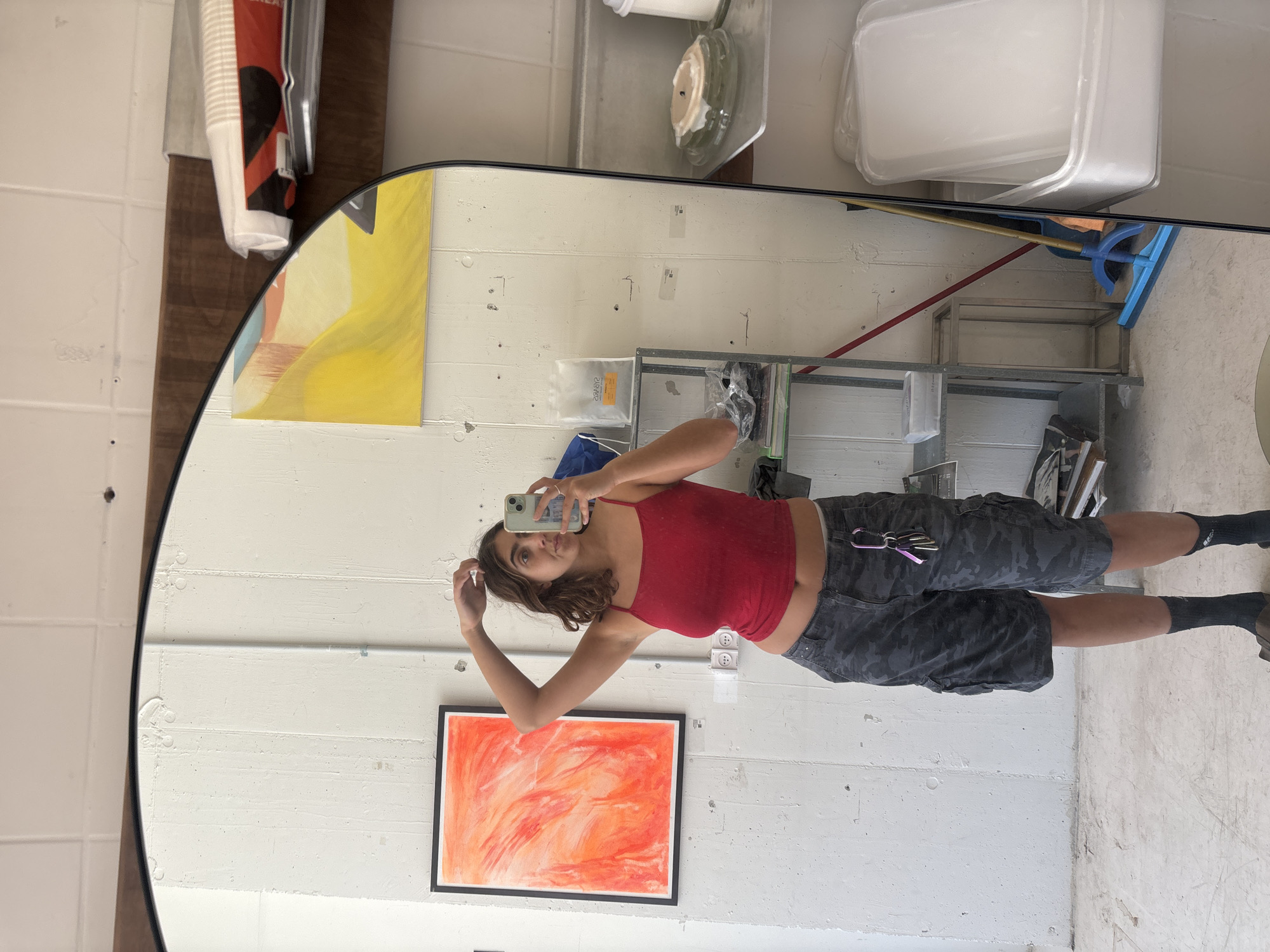 The image is a reflection in a mirror. A woman is holding a phone, presumably taking the picture of her reflection. She is wearing a red tank top and patterned shorts, and has dark hair. She stands in what appears to be an indoor space with white concrete walls. A painting with orange and red hues in a black frame is visible on the wall behind her. There's also a yellow painting in the upper left of the mirror. Above the woman, a shelf and a broom are partially visible.
There is no information available to verify whether the image was taken in Tel-Aviv, Israel.