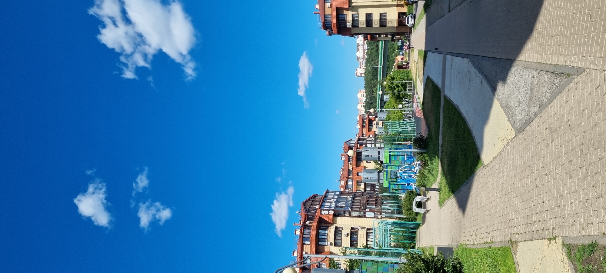 The image depicts a residential area with apartment buildings and a playground, located in Romashkovo, Russia. The buildings are multi-story with red tiled roofs and light-colored facades, some featuring balconies and modern windows. A paved walkway with brickwork runs through the foreground, leading towards a playground area equipped with colorful structures and green fencing. Adjacent to the buildings are patches of grass and trees. In the background, a bridge spans across a green landscape, with more buildings visible in the distance. The sky is a clear blue with scattered white clouds, indicating a sunny day. A few individuals are visible walking in the distance, and a car is parked on the right side of the walkway.