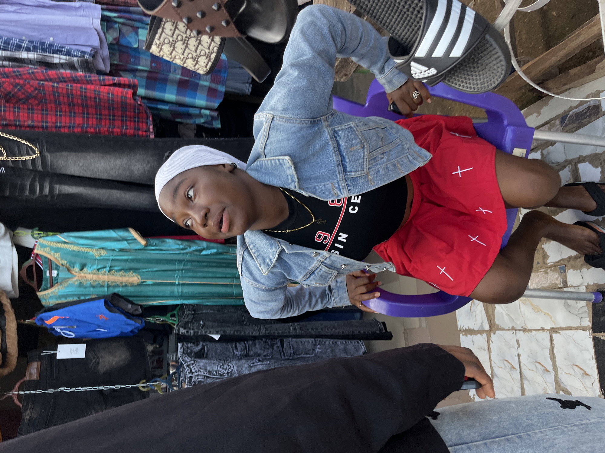 A young Black woman wearing a white headscarf, a denim jacket, and a red skirt with white cross patterns is seated on a purple plastic chair in front of a clothing stall in Bukuru, Nigeria. She is looking upwards with her mouth slightly open. A person's torso and arm, wearing a black t-shirt, are visible on the left, and this person appears to be holding a phone. Behind the woman, various garments are displayed, including black jeans, blue shorts, a teal-colored kaftan with gold trim, and plaid shirts. To the right of the woman, a pair of Adidas slide sandals is visible. The stall appears to be located outdoors, with tiled flooring visible beneath the chair.