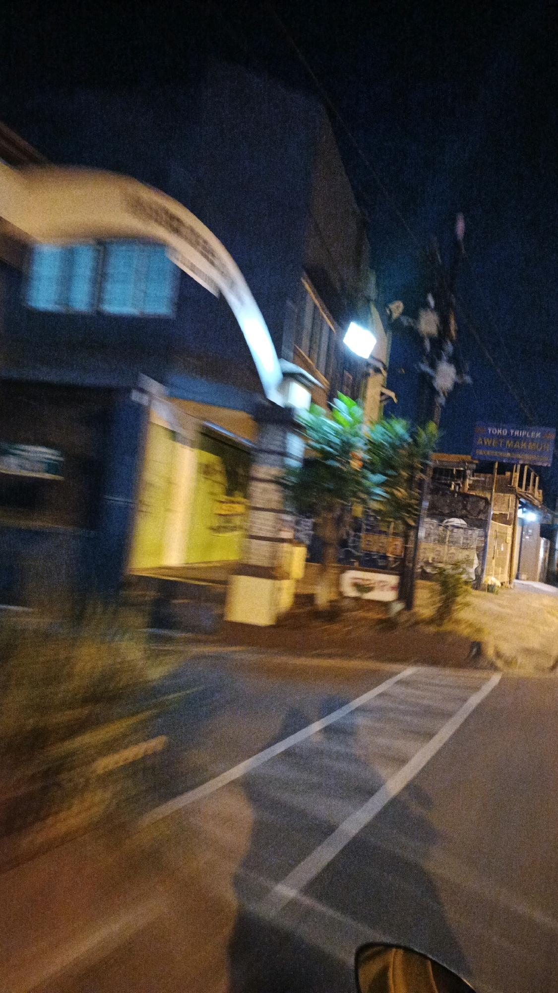 A nighttime, motion-blurred, street-level view captures a scene in Surakarta, Indonesia. A building with a large, arching light fixture extends across the top of the frame. The building appears to have windows with a blue hue and a yellow entrance or display. A street sign, partially obscured, reads "TOKO TRIPLEK AWET MAKMUR". Trees and power lines are visible in front of the building, with a street lamp casting light. The foreground shows a street with white lane markings and a prominent shadow of what appears to be the photographer and their vehicle. The overall impression is of a transient moment captured while in motion.