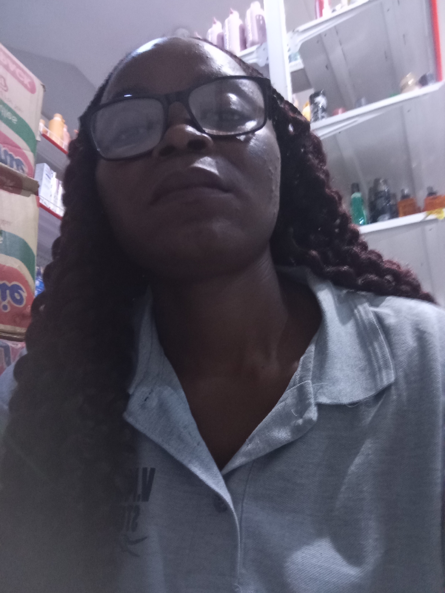A close-up selfie captures a woman from the chest up, her face tilted slightly upwards. She is wearing black-rimmed glasses and her dark, braided hair cascades down her shoulders. The woman is dressed in a light gray polo shirt with a collar. On the left side of the shirt, a dark emblem with text is visible; some of the visible characters are "M," "V," and "12". The background is slightly blurred, revealing shelves stocked with various products, including bottles and boxes. The overall lighting suggests an indoor setting, possibly a store or a home with shelves arranged for display. The woman's expression appears neutral or contemplative. The location context is Howayi, Nigeria.