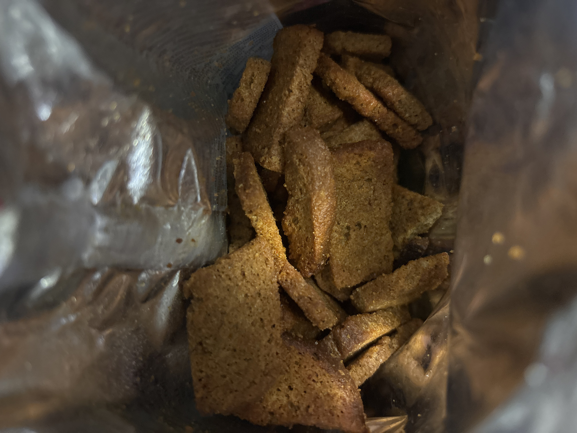 A pile of toasted rye croutons is visible inside a shiny, silver bag. The croutons are rectangular and irregular in shape, with some broken into smaller pieces. They appear to be seasoned with spices, giving them a slightly reddish-brown hue. The bag is partially open, allowing a view into the contents. The background is blurry, suggesting a shallow depth of field. The image is lit from above, casting shadows on the croutons. The location is Unknown, Unknown.