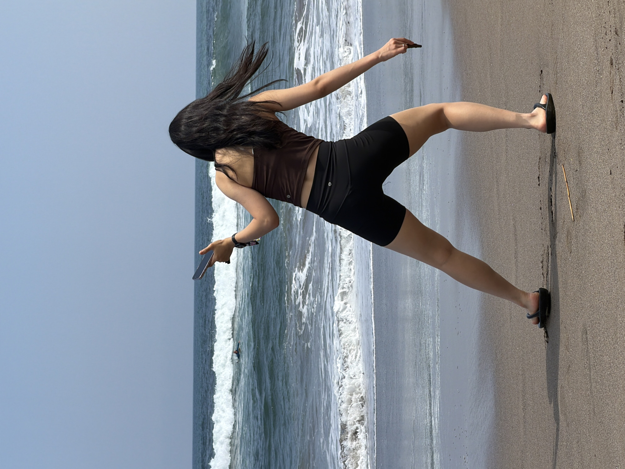 A person is standing on a sandy beach in North Kuta, Indonesia, with the ocean and sky in the background. The person is facing away from the camera, wearing a brown tank top and black shorts. They are holding a smartphone in their right hand, extended forward as if taking a photo or video. Their left arm is bent, and they are holding what appears to be a small object in their left hand. There are waves in the ocean, and the sky is clear and blue. The sand is a dark brown color, and there is a small stick on the beach near the person's feet.